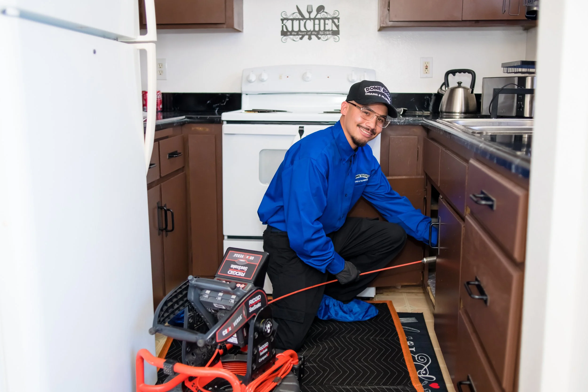 A technician in a blue shirt and black gloves kneels on the floor of a kitchen, working on the cabinet beneath the sink. The man is smiling and wearing glasses and a black cap. There is a piece of electronic testing equipment on the floor, connected with orange cables. The kitchen has brown cabinets, a white stove, and a stainless steel kettle on the counter.