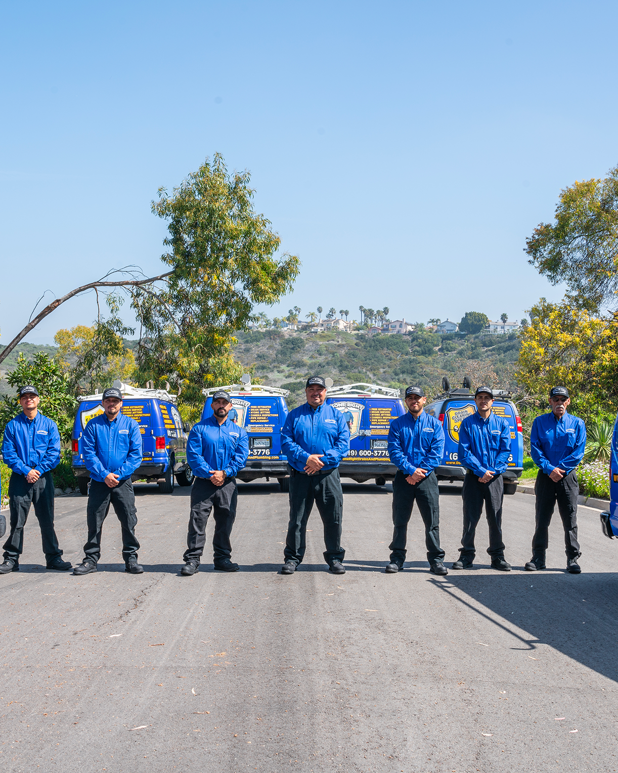 Seven servicemen in blue uniforms standing in front of service vans on a sunny day with trees and hills in the background.