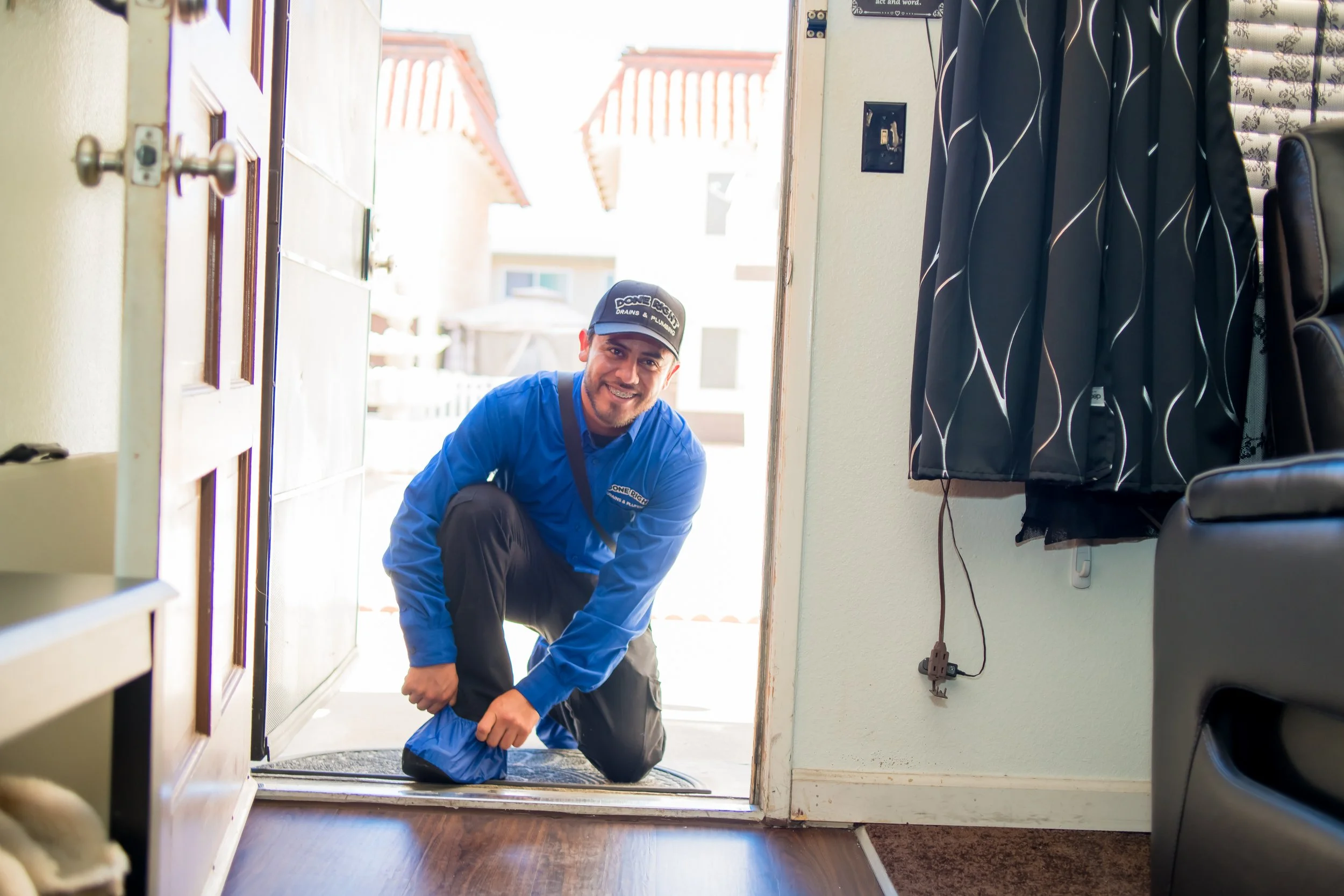 A smiling man wearing a uniform kneels at the door indoors, with sunlight visible outside. He wears a black cap and a blue shirt with a logo, and appears to be working or greeting.