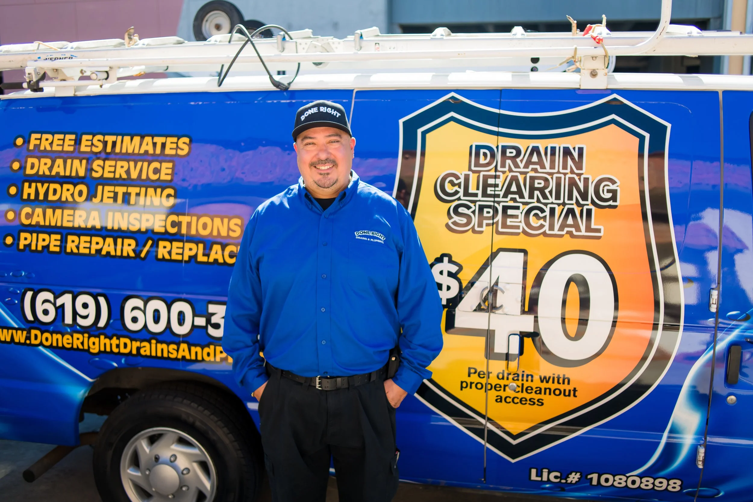 A man wearing a blue shirt and a black cap standing in front of a blue van advertising drain cleaning services, with the company logo and a list of services.