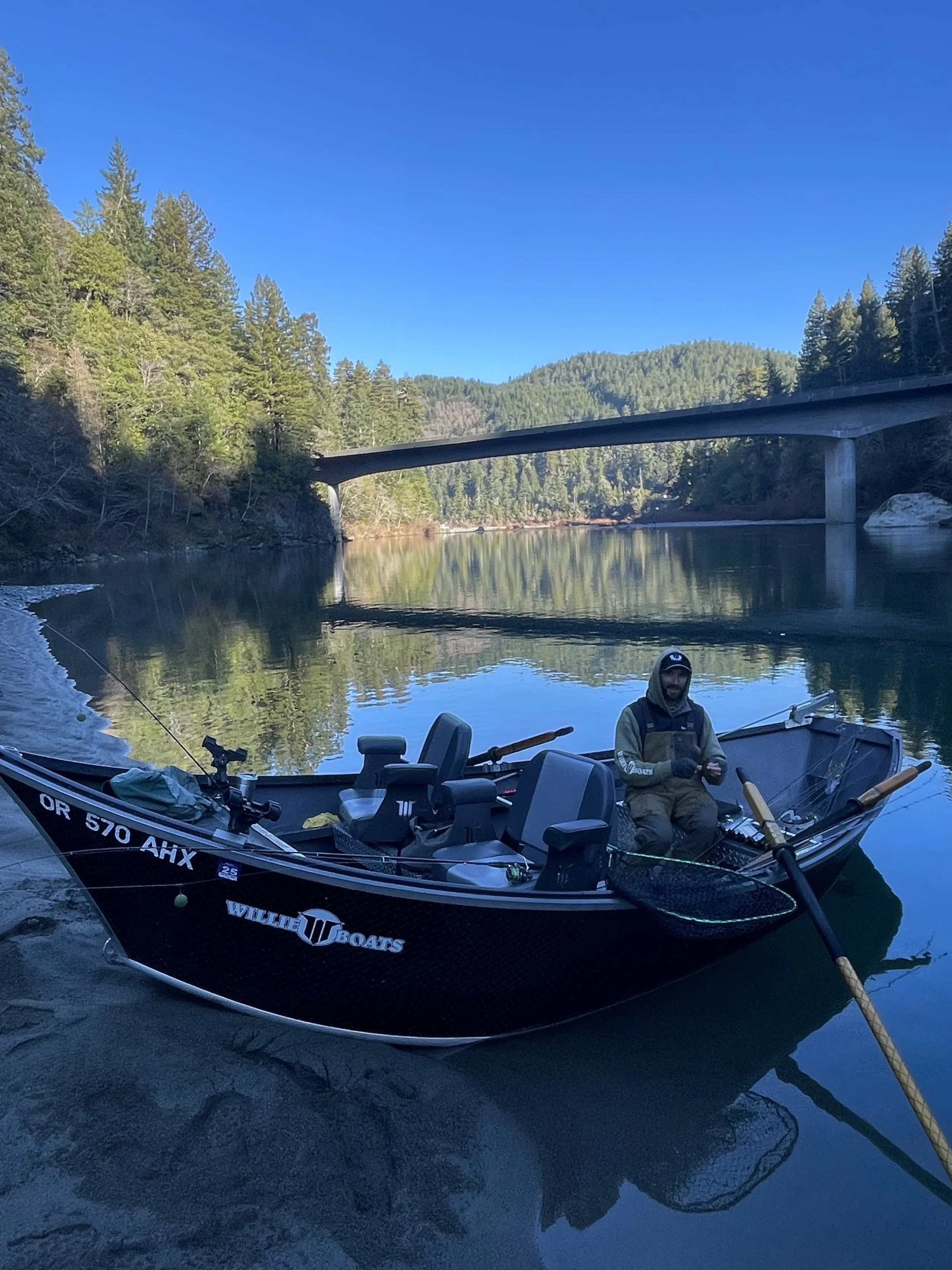 A man sitting in a boat on a calm river with lush green trees on either side and a bridge crossing overhead. The boat is equipped with fishing gear and the man is wearing fishing attire.