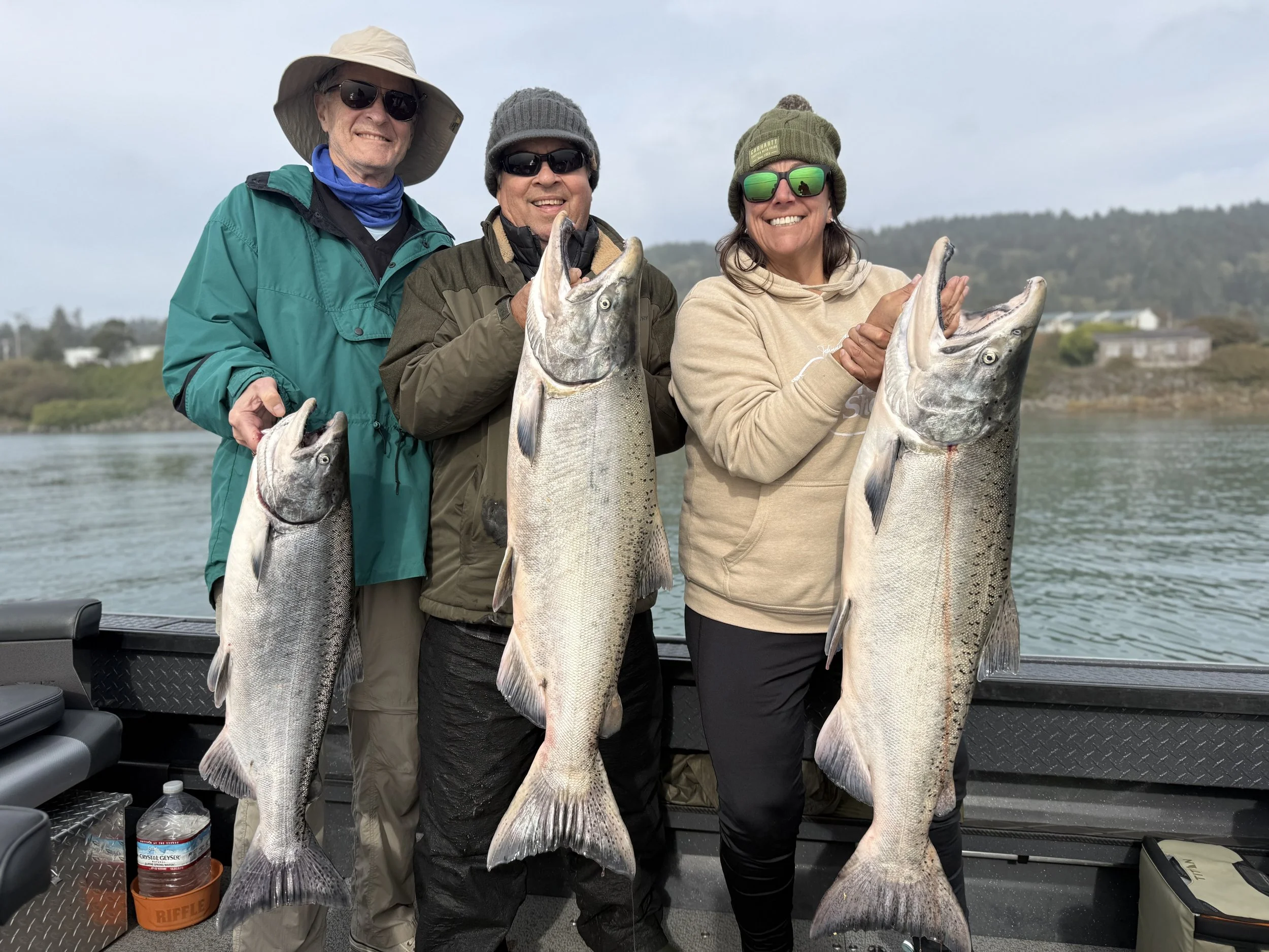 Three people on a boat holding large fish they caught, with water and hills in the background.