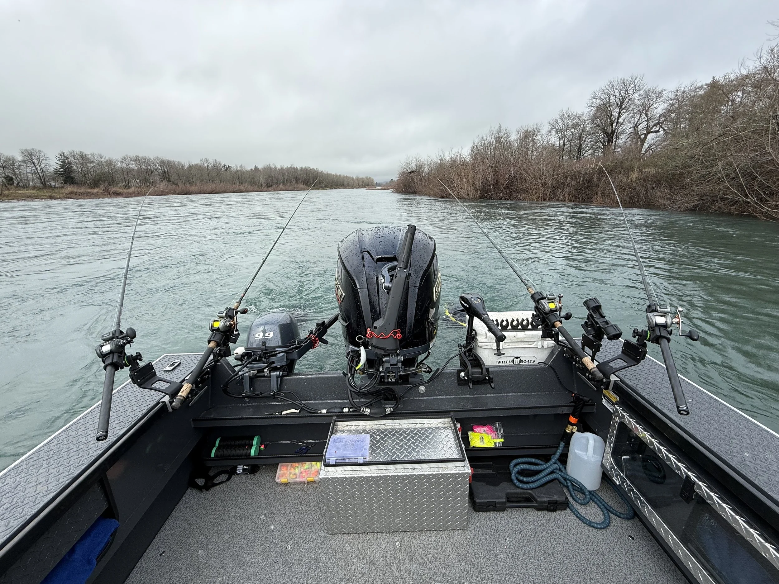 View from the front of a boat on a calm river with fishing poles on each side and a gray overcast sky.