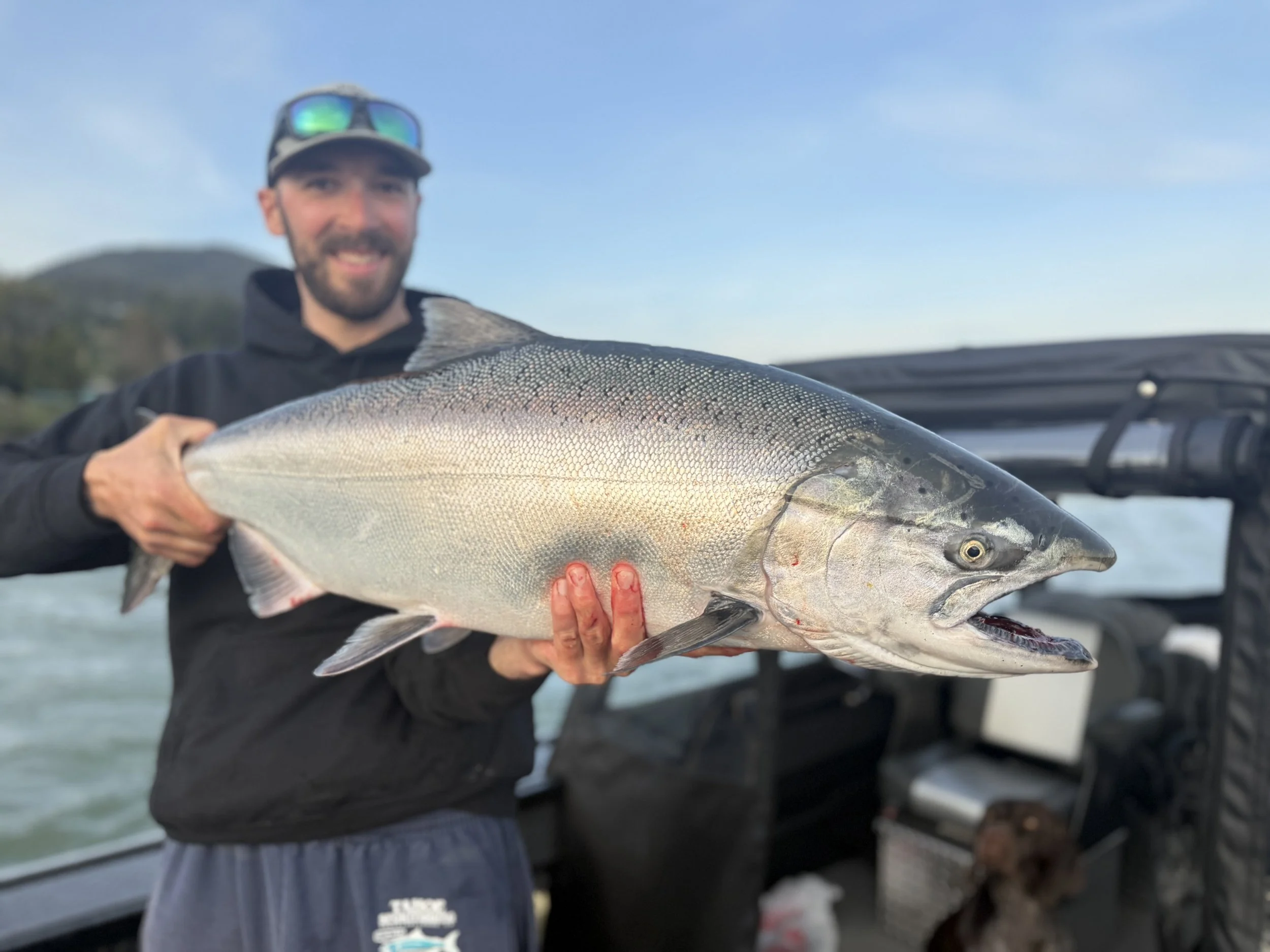 Man holding a large fish on a boat with water and distant landscape in the background.