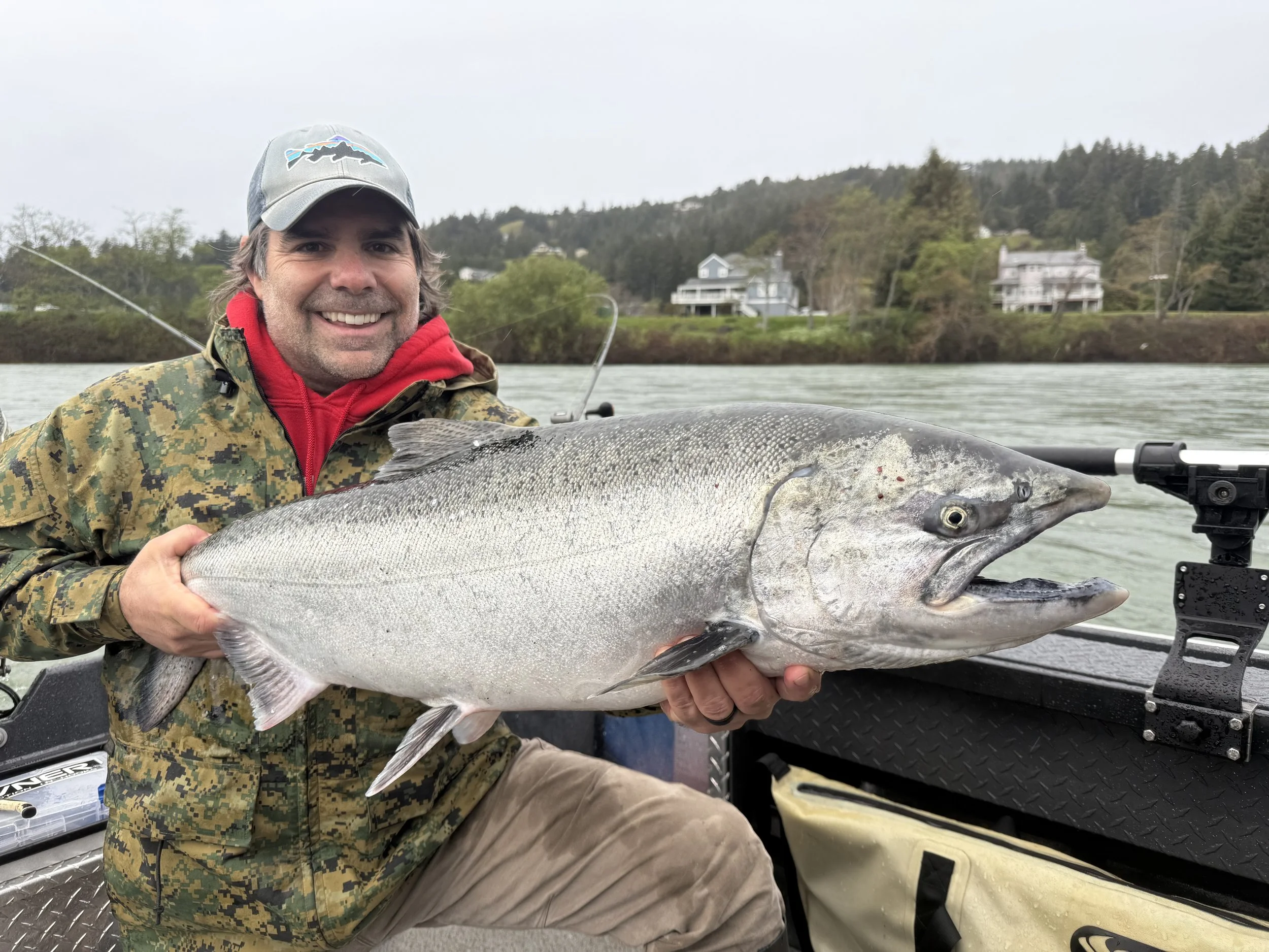 Man in camouflage jacket and red hoodie smiling while holding a large fish on a boat with a lake and houses in the background.