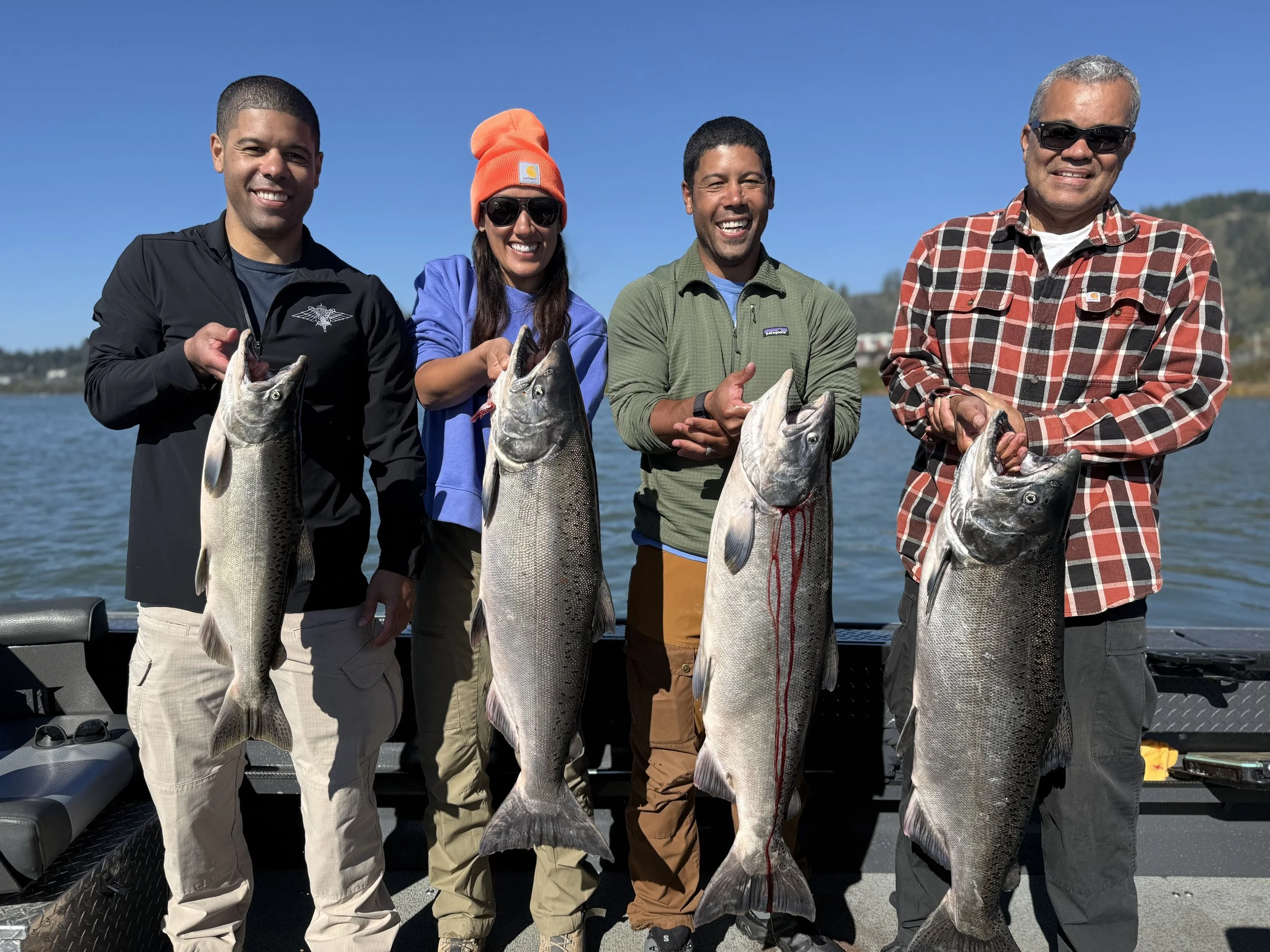 Four people on a boat holding large fish they caught, with water and distant shoreline in the background.