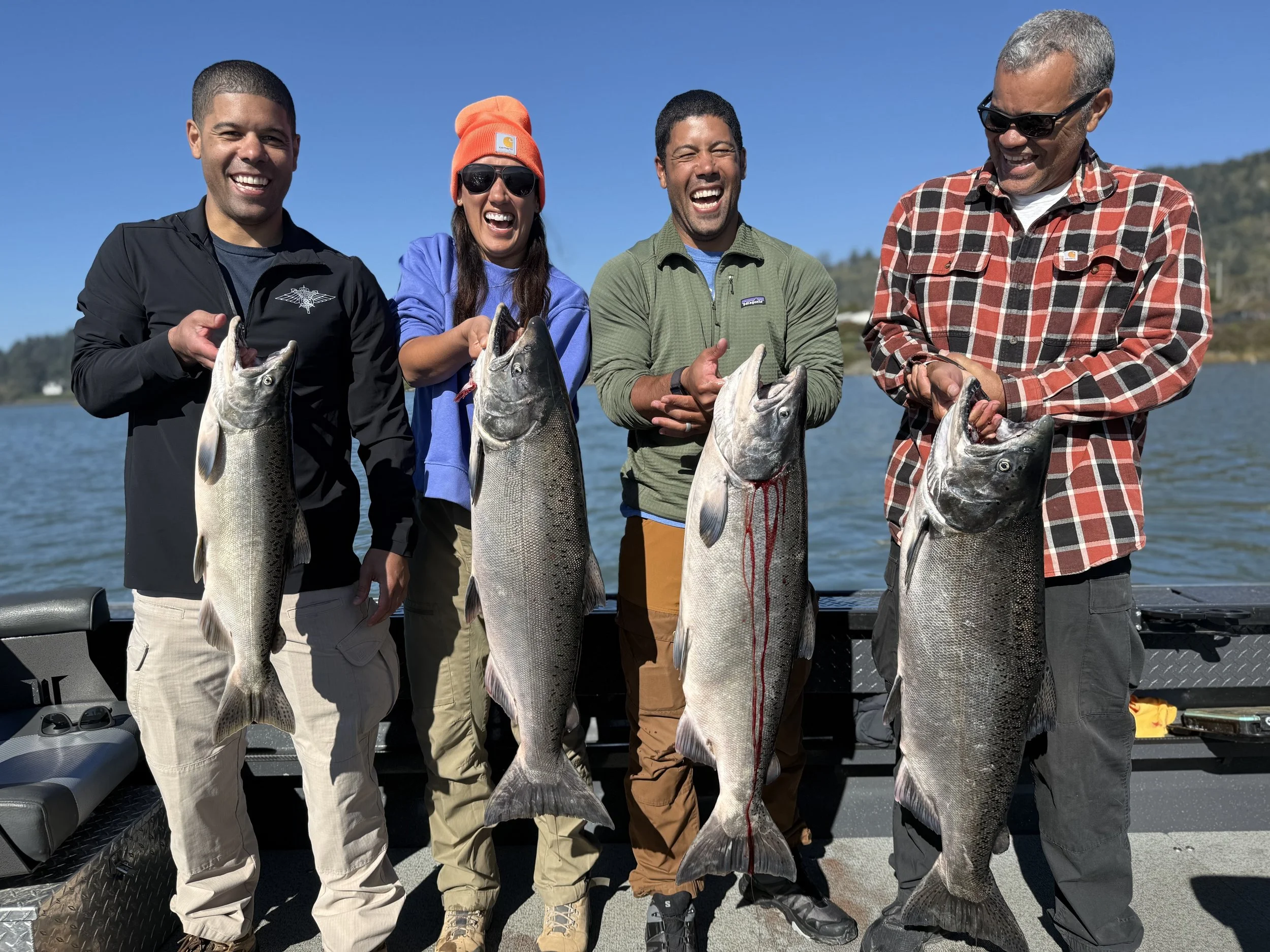 Four people standing on a boat, each holding a large fish, smiling after a successful fishing trip on a lake with mountains in the background.