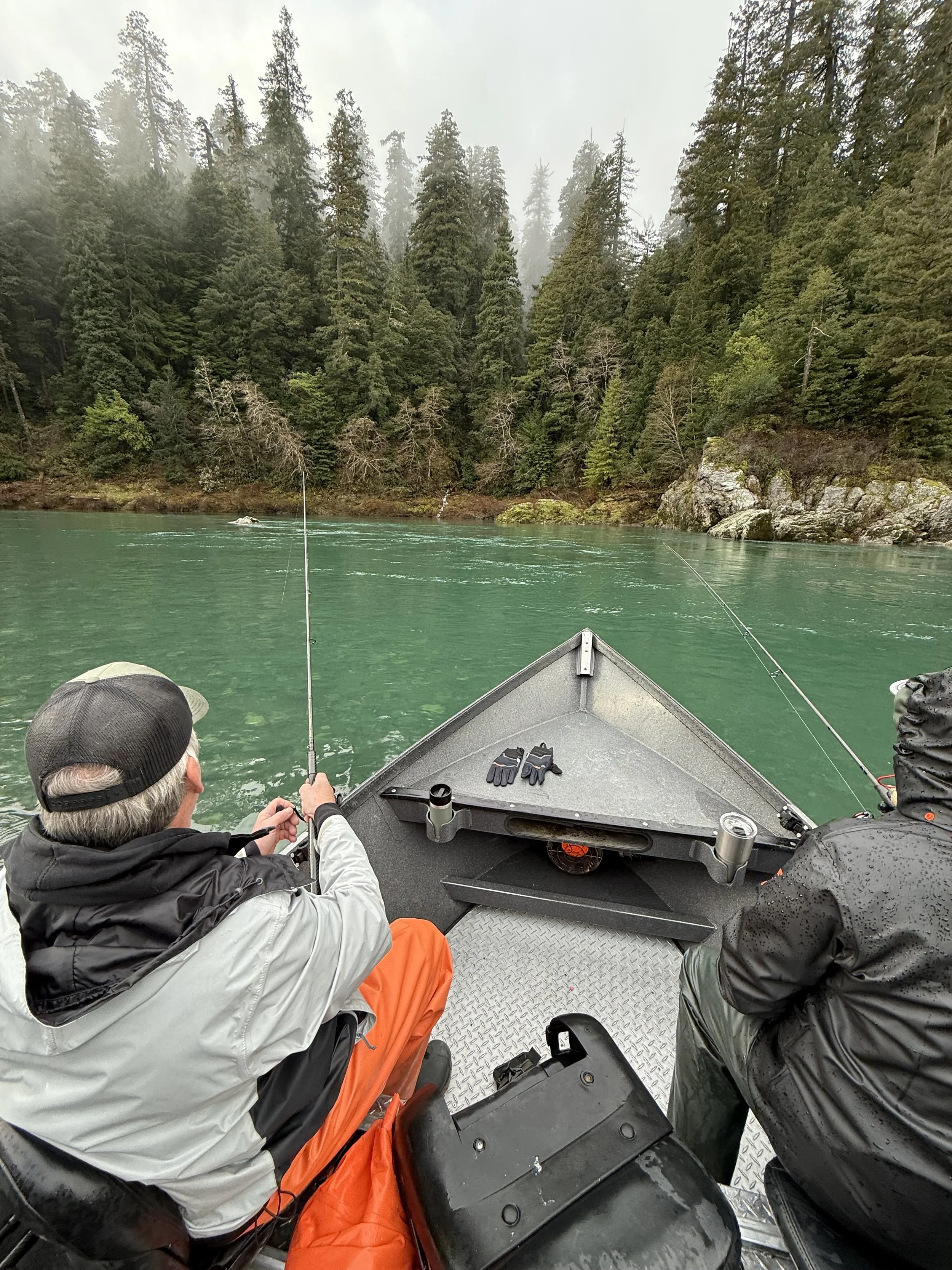 Two men fishing from a boat on a river surrounded by forested mountains in rainy weather.