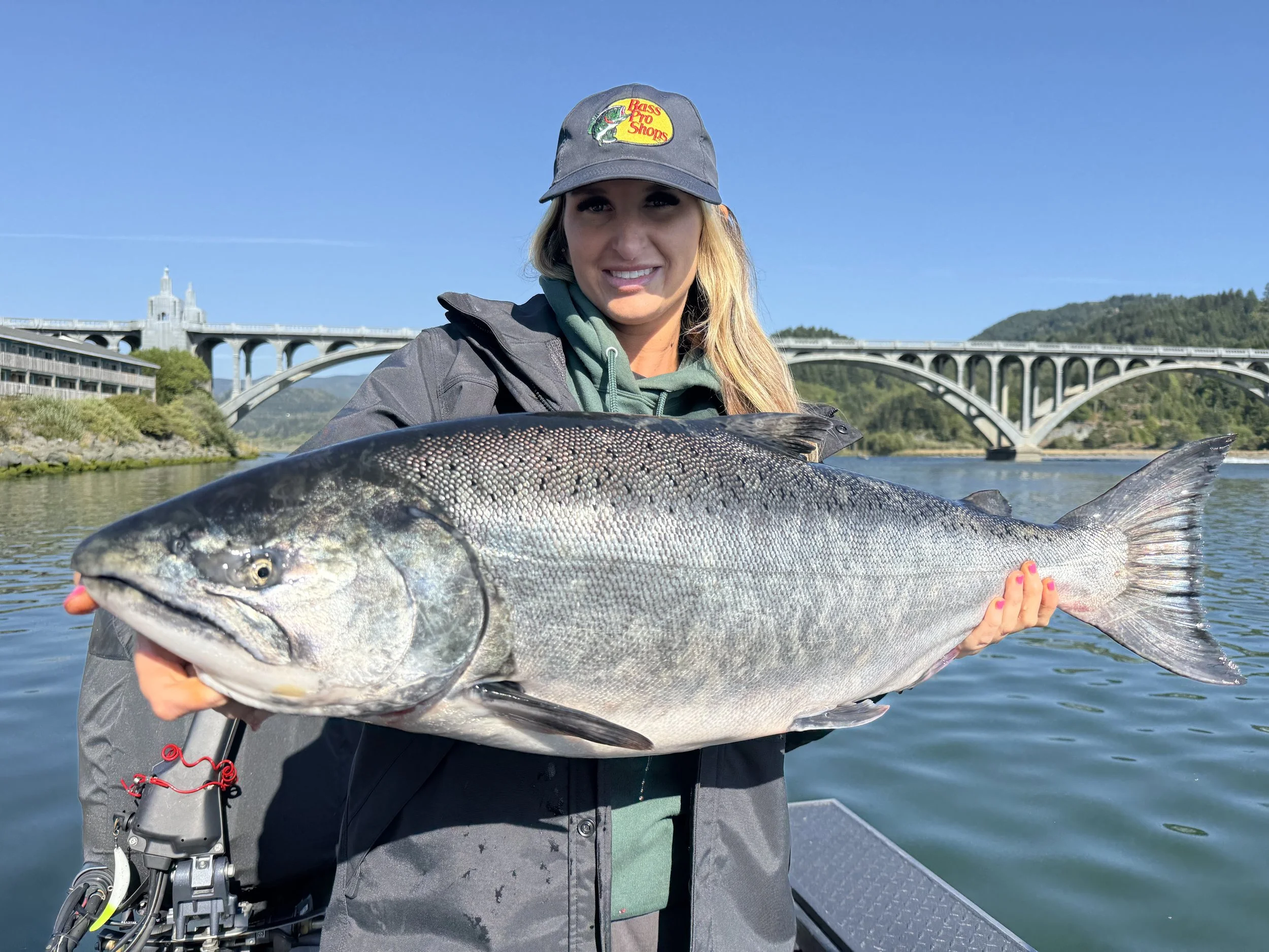 A smiling woman holding a large fish with water and a bridge in the background.