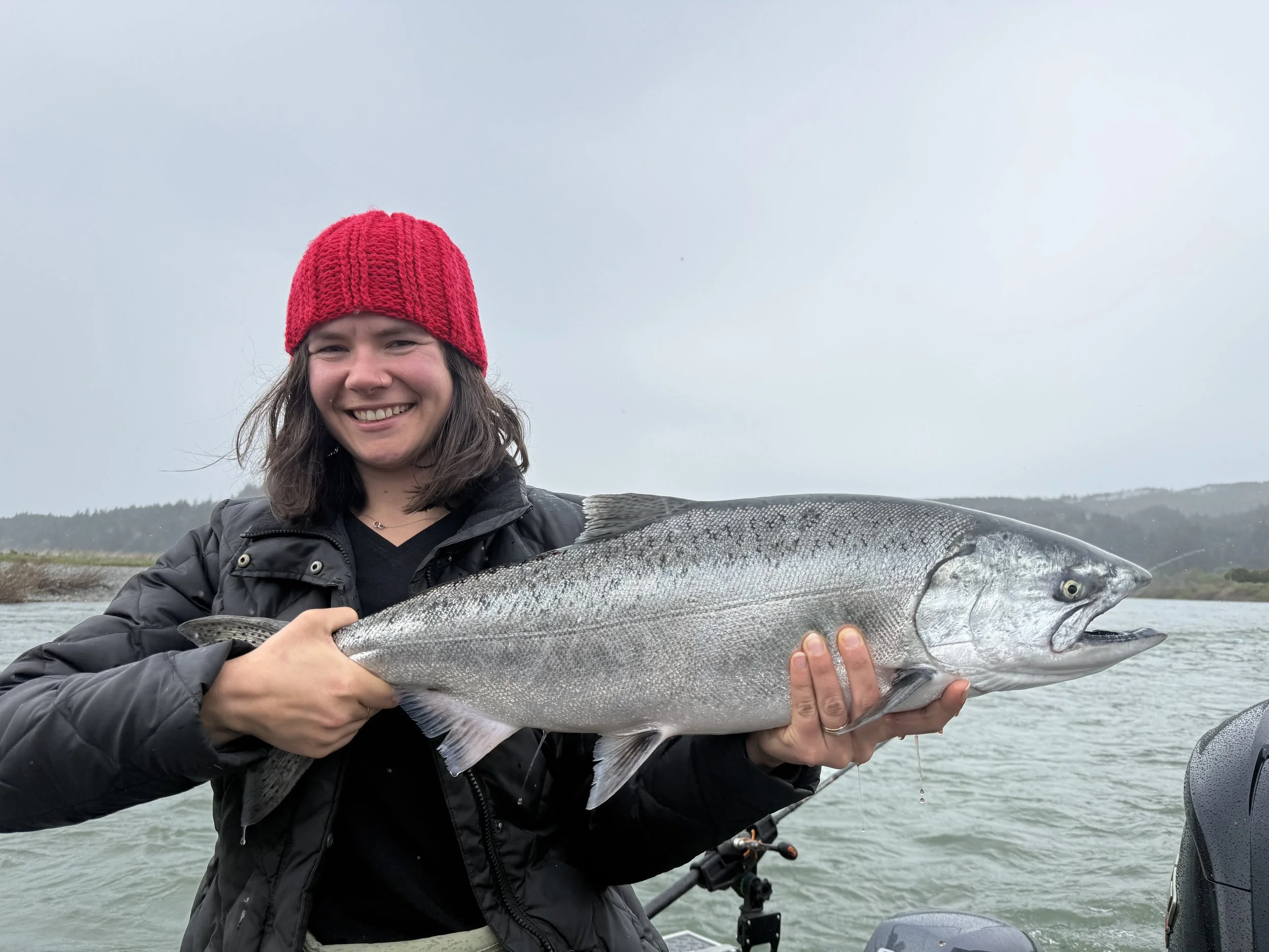A smiling woman wearing a red knit hat and black jacket holds a large silver fish with a wide-open mouth on a boat in a body of water, with a cloudy sky and distant hills in the background.