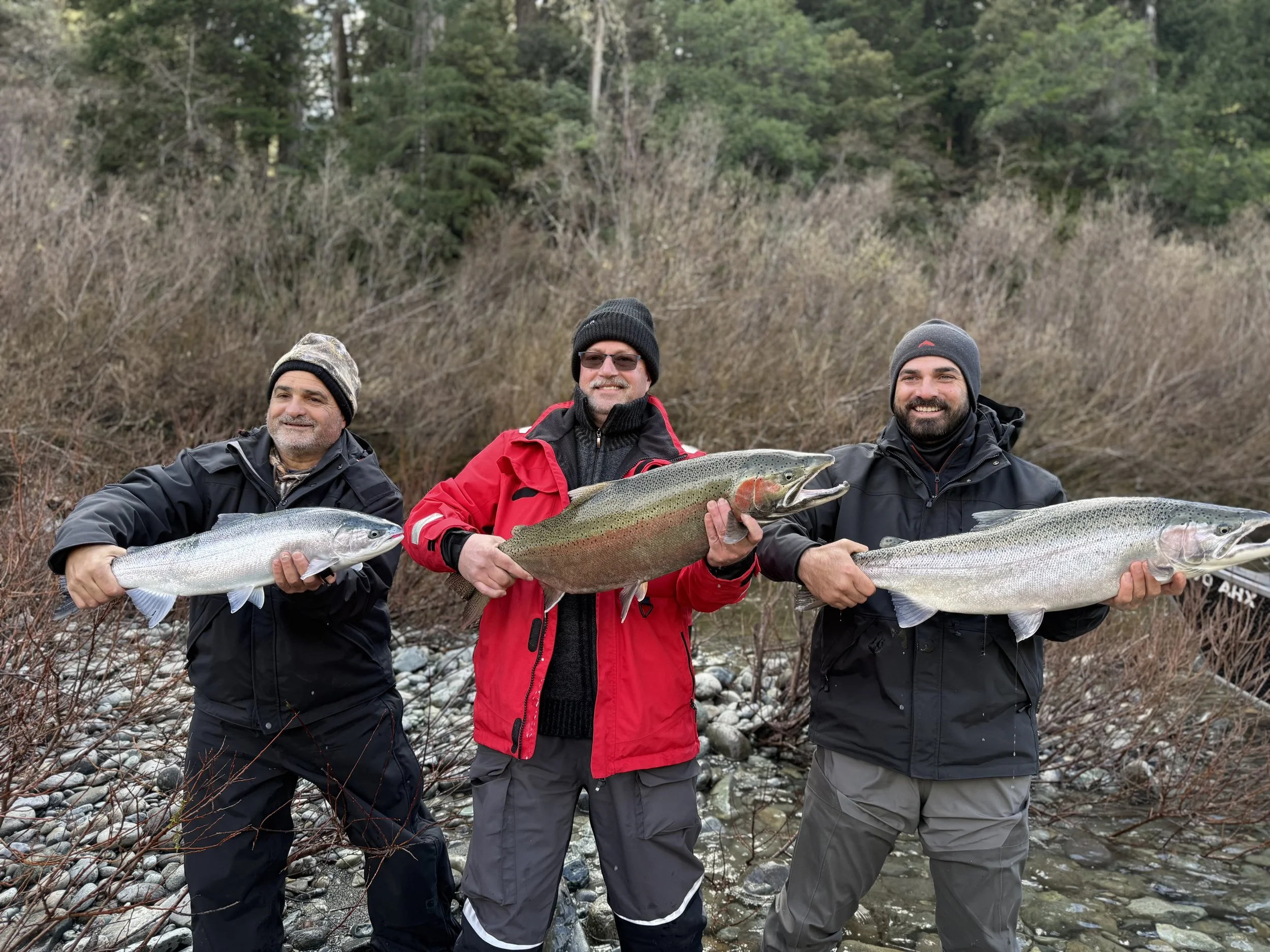 Three men holding large fish outdoors in a natural setting with trees and rocks in the background.