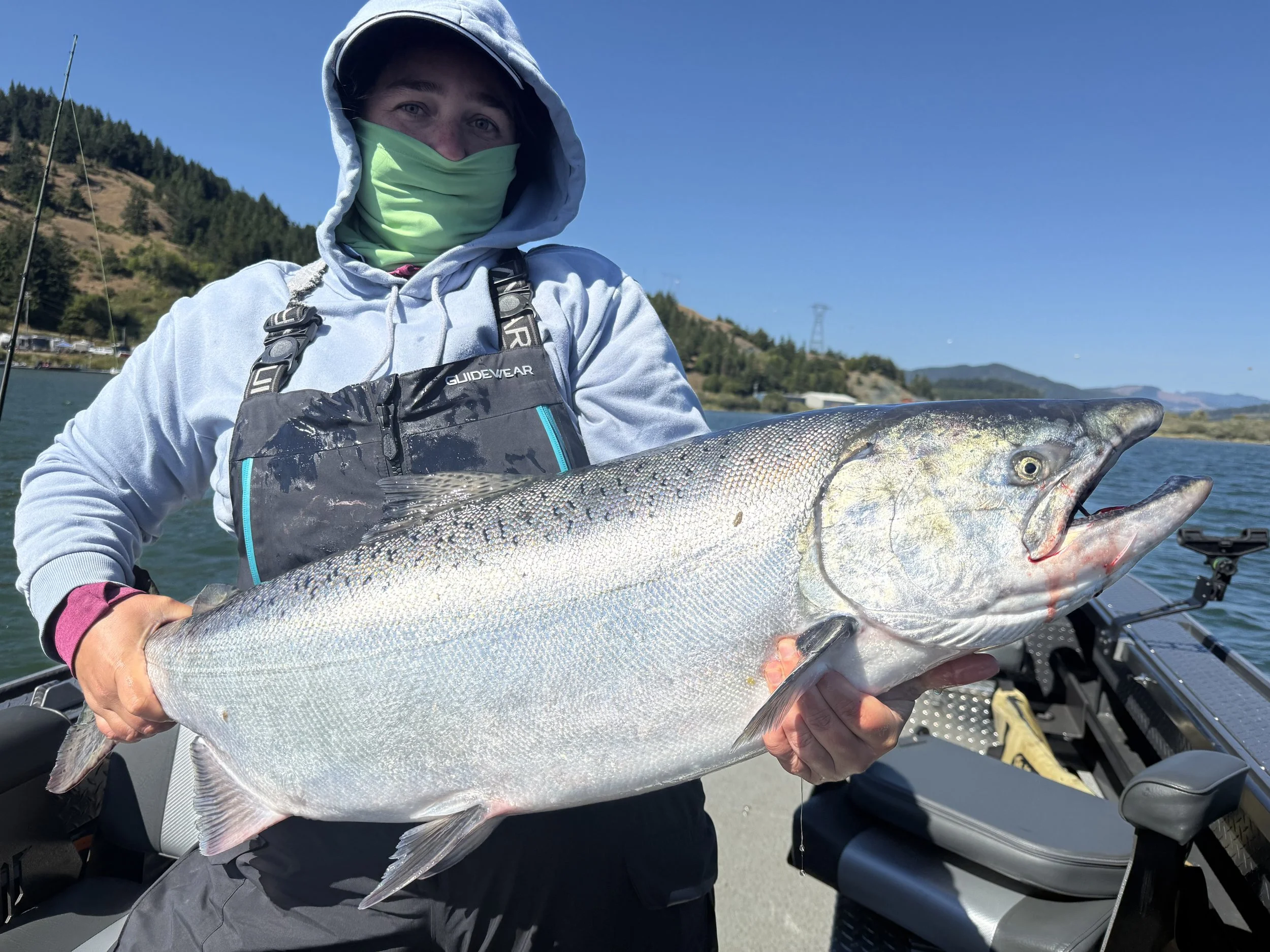 Person in fishing gear holding a large fish on a boat with a scenic water and mountain background.