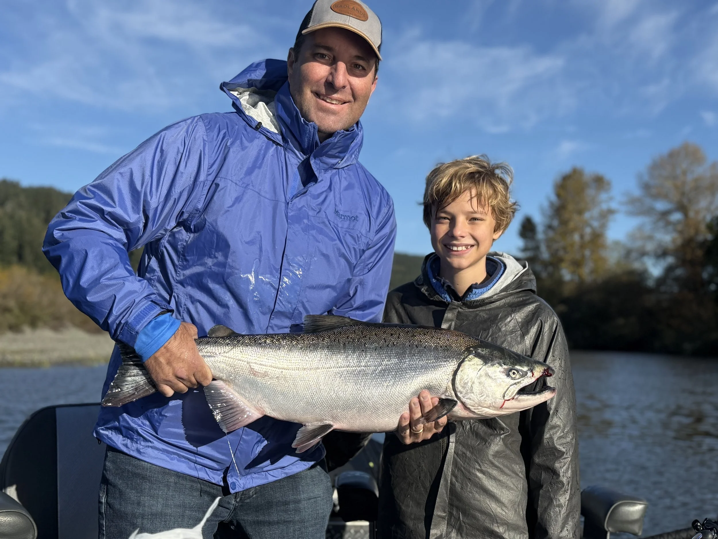 A man and a boy standing outdoors on a boat, smiling at the camera, holding a large fish between them with a body of water and trees in the background.