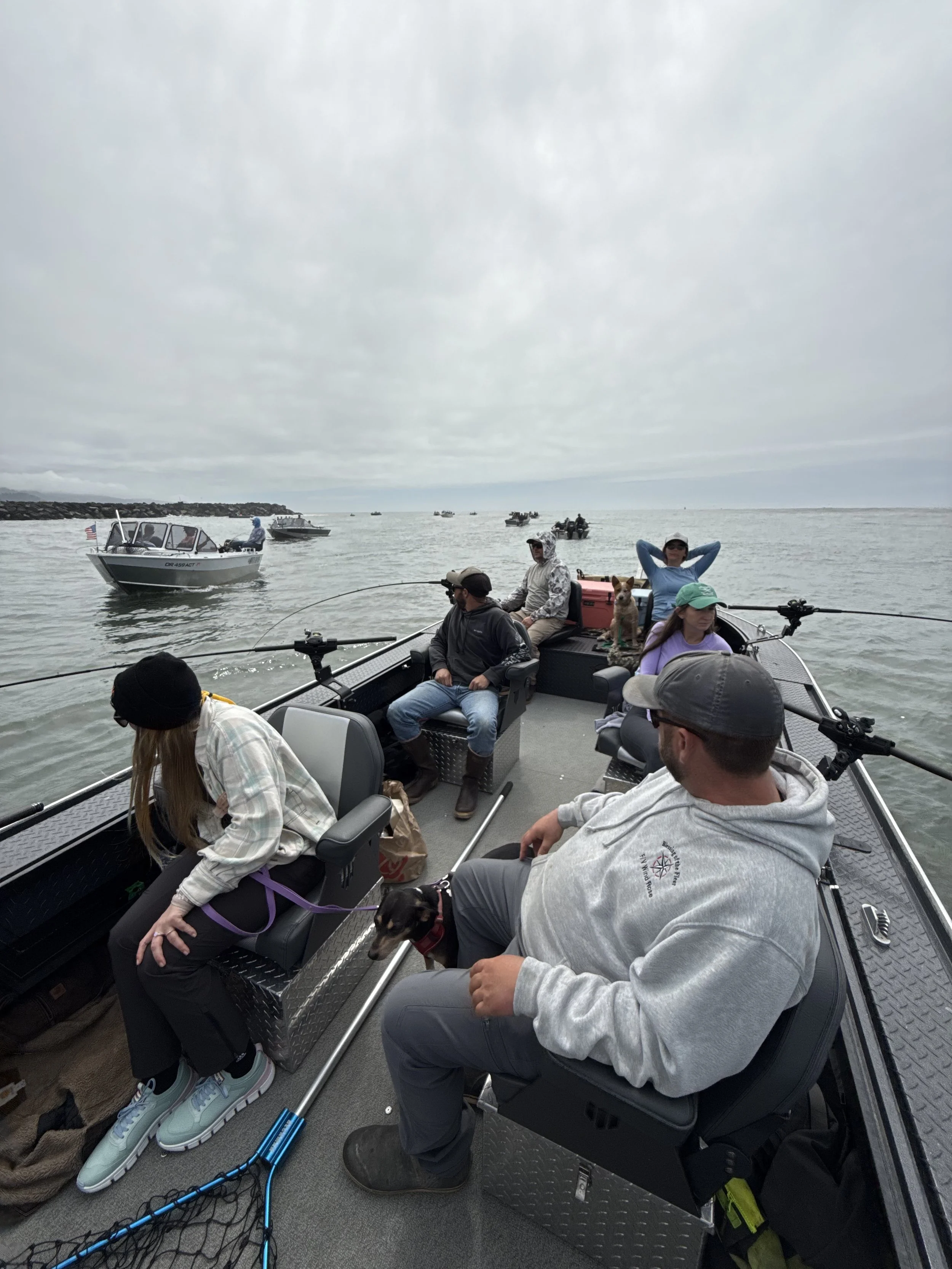 A group of people on a boat enjoying a day of fishing, with some sitting, some standing, and dogs present on board, with other boats visible in the water under a cloudy sky.