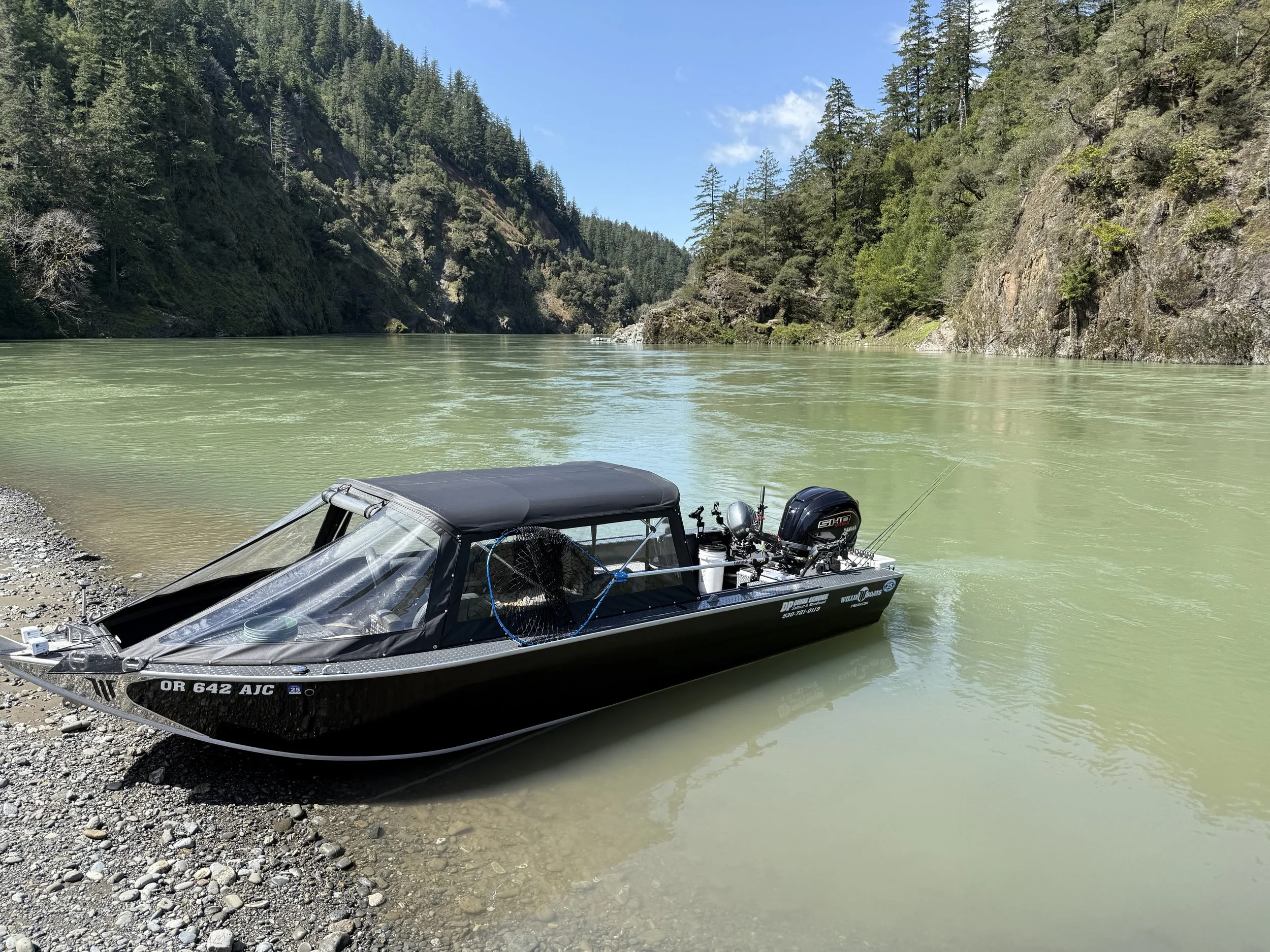 A small black and silver motorboat is docked on the rocky shore of a greenish river, surrounded by forested hills under a partly cloudy sky.