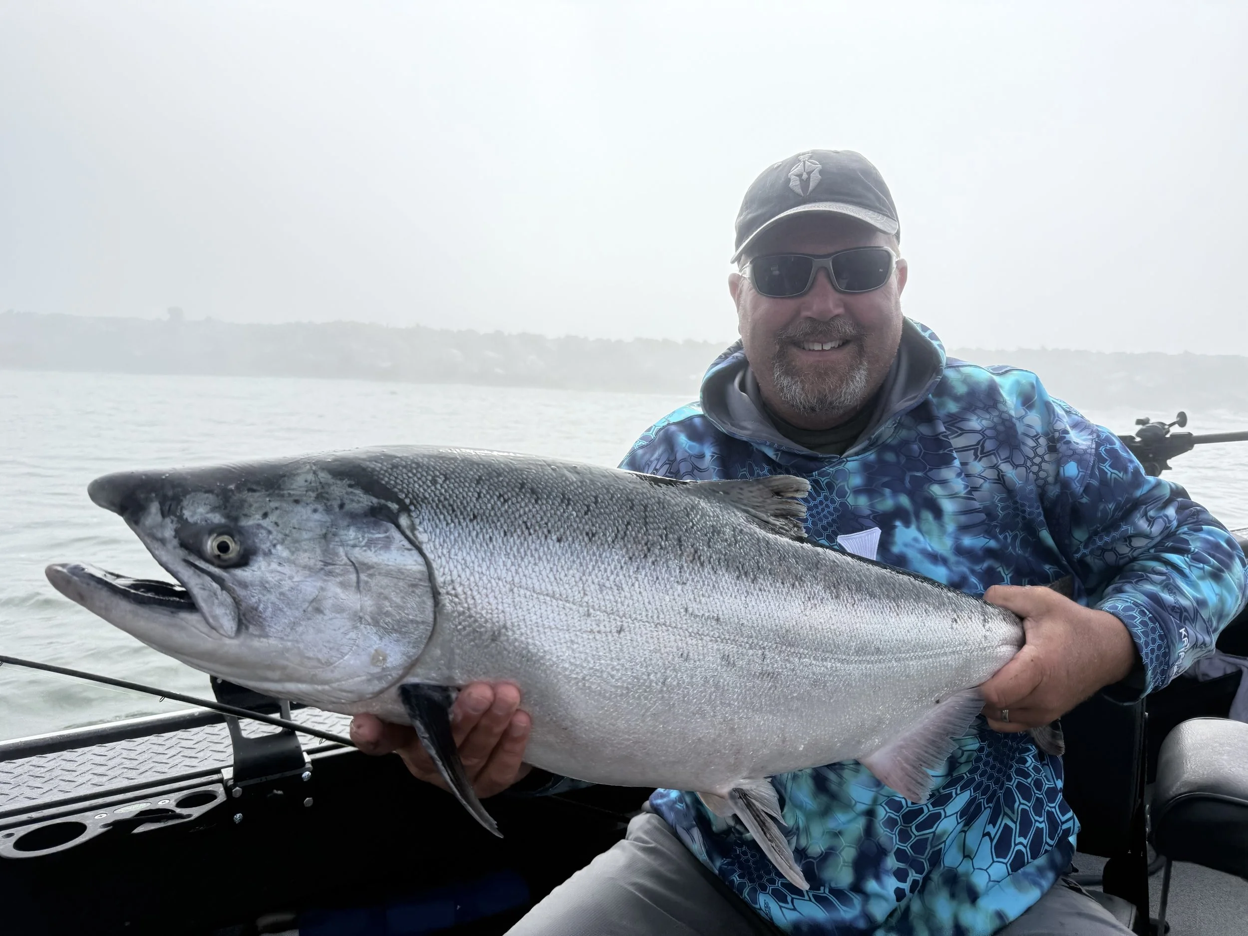 A man wearing sunglasses, a cap, and a blue patterned jacket smiling and holding a large fish on a boat during daytime.