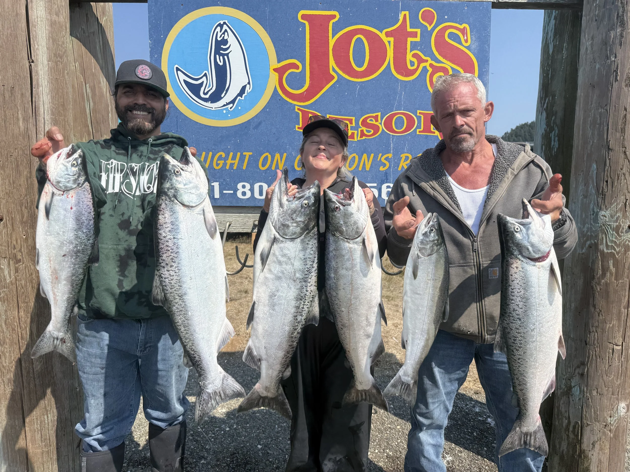 Three people standing outdoors holding large fish in front of a sign that reads "Jots Bison" with a fish logo. The man on the left has a beard, wearing a cap and hoodie. The woman in the middle is wearing a cap and black jacket, and the man on the ri