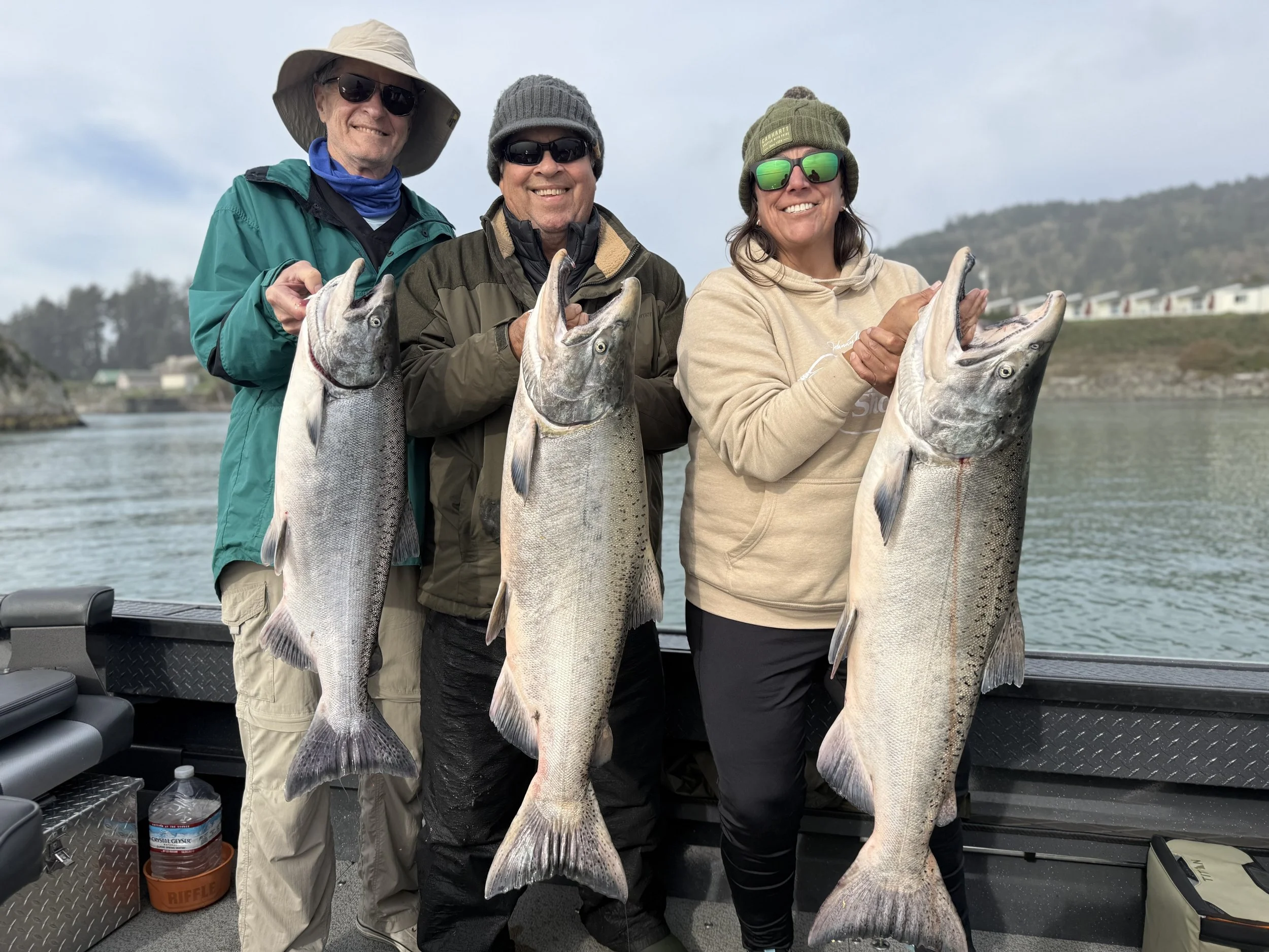 Three people standing on a boat holding large fish, smiling. They wear outdoor gear, hats, and sunglasses, with a body of water and hills in the background.