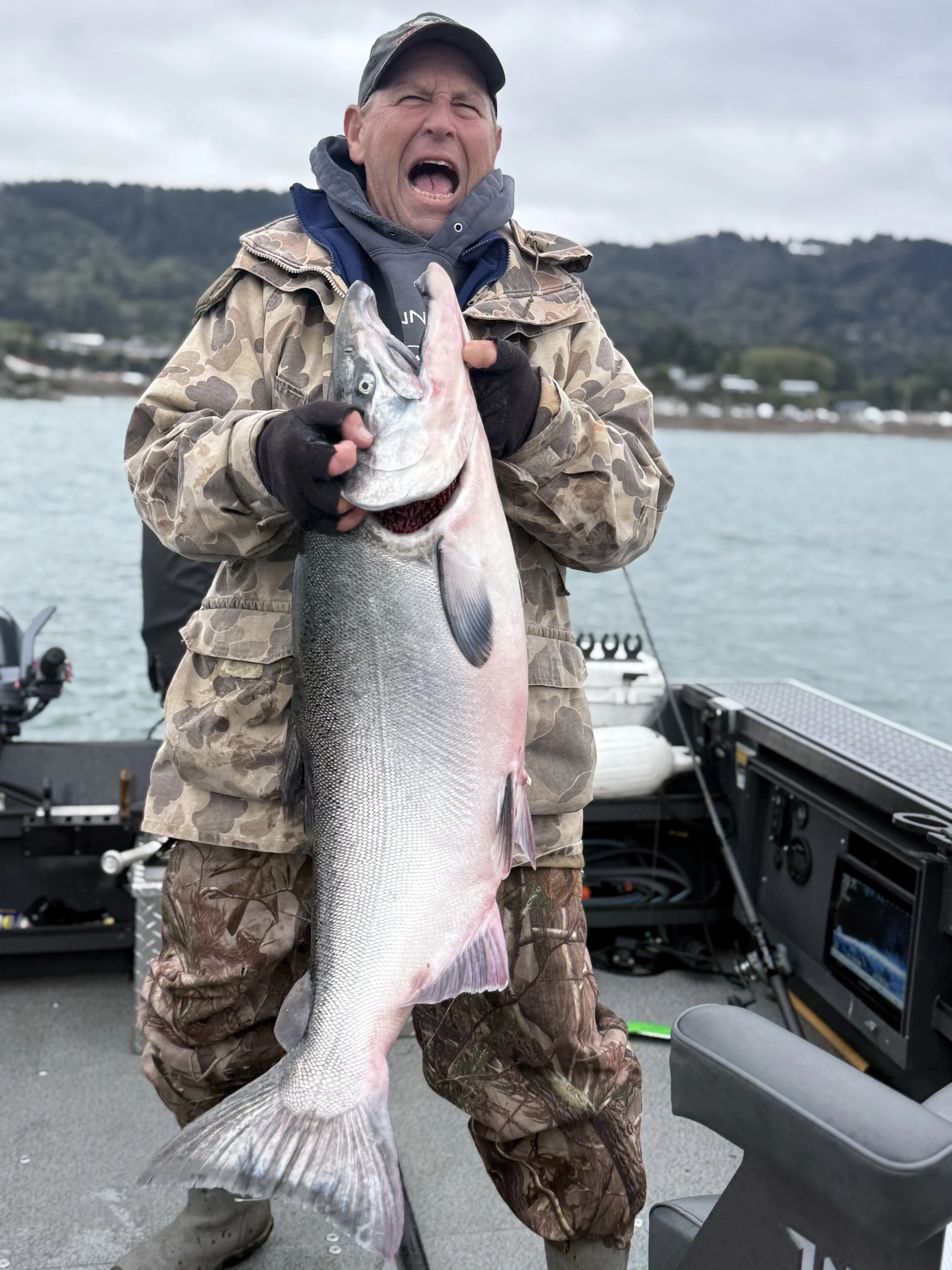A man in camouflage jacket and gloves holding a large fish on a boat, with water and hills in the background.