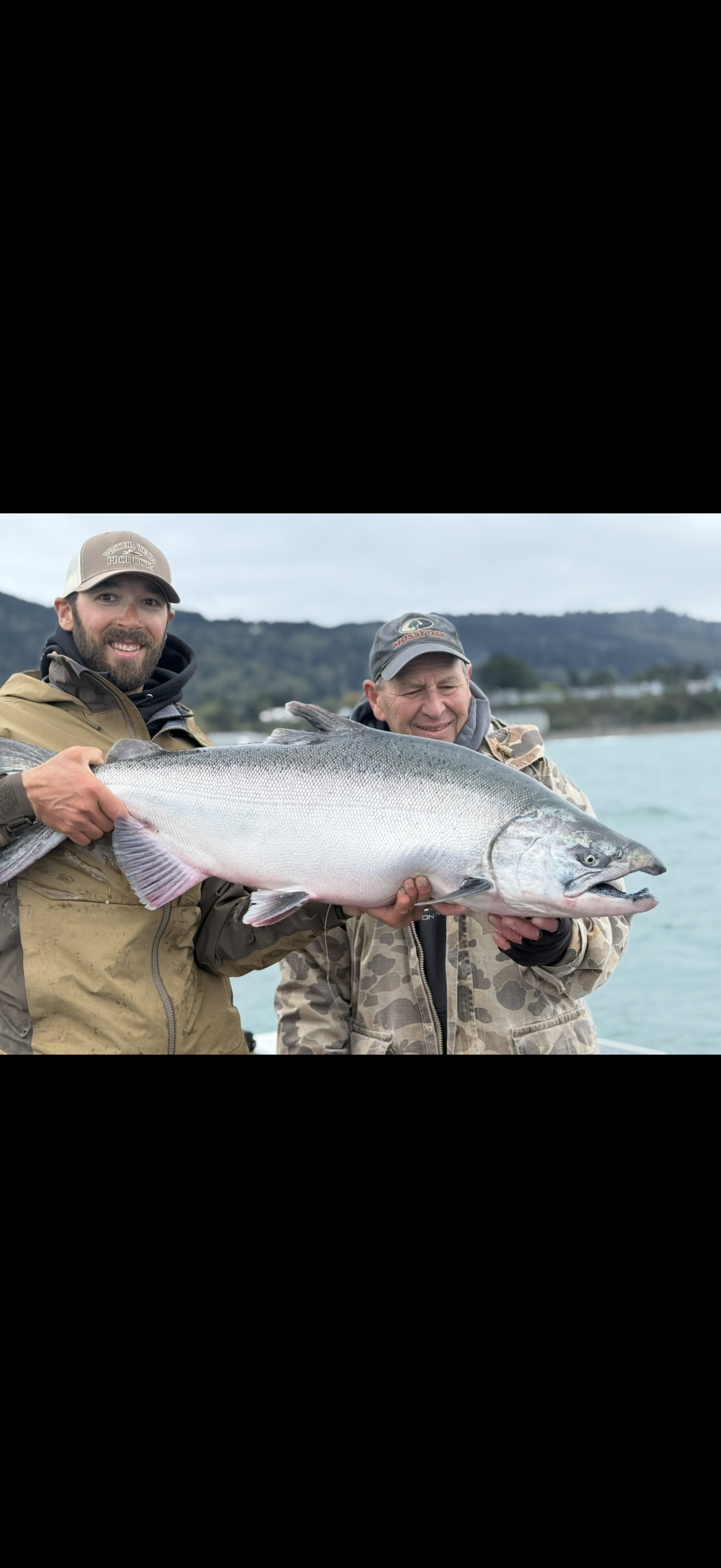 Two men holding a large fish, with a lake and hills in the background, and a black rectangle in the upper right corner with a phone number and call to action.