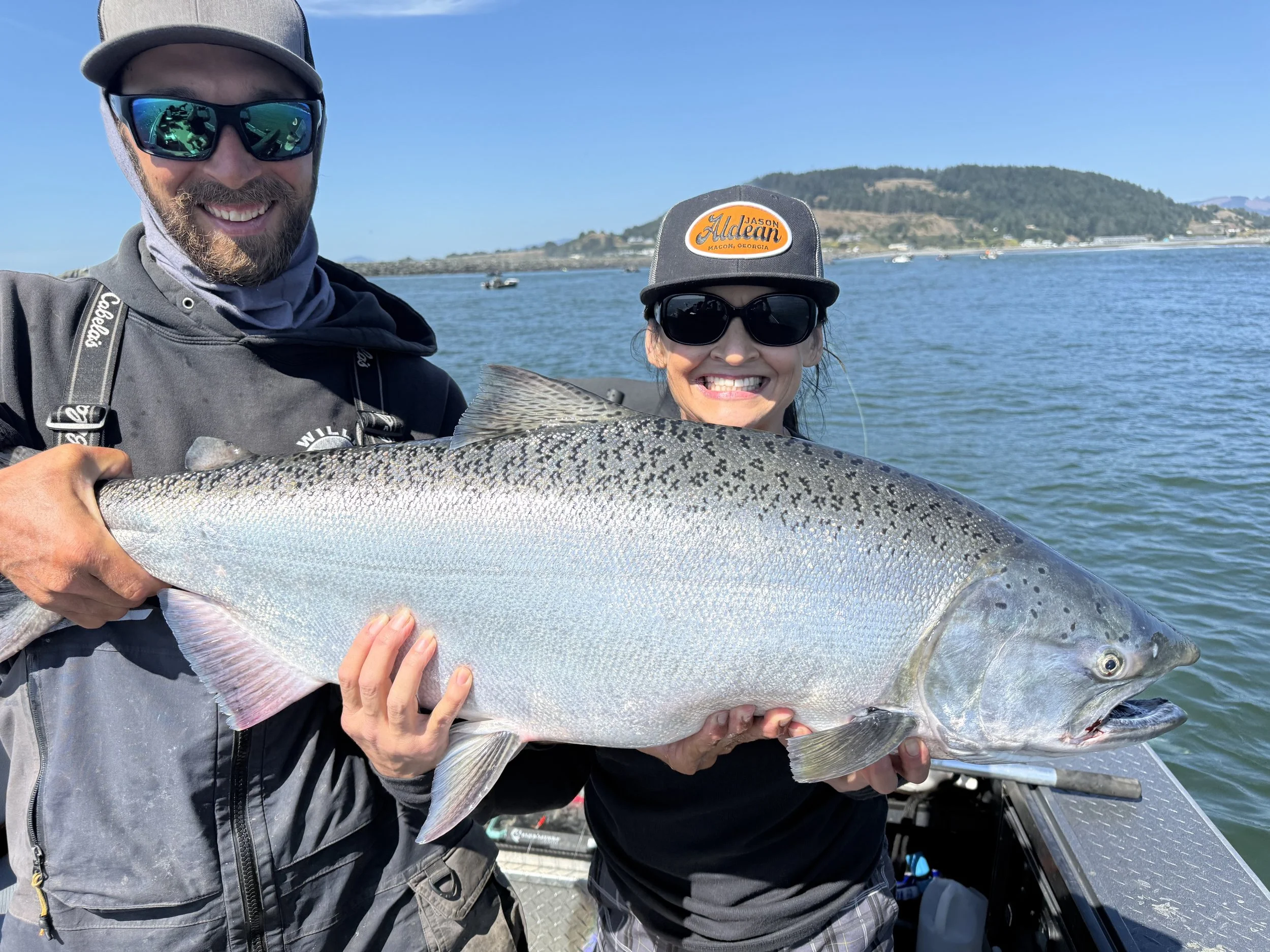 Two people on a boat holding a large fish with a scenic water and mountain background. The man on the left is wearing sunglasses and a jacket, and the woman on the right is wearing a cap and sunglasses, both smiling.