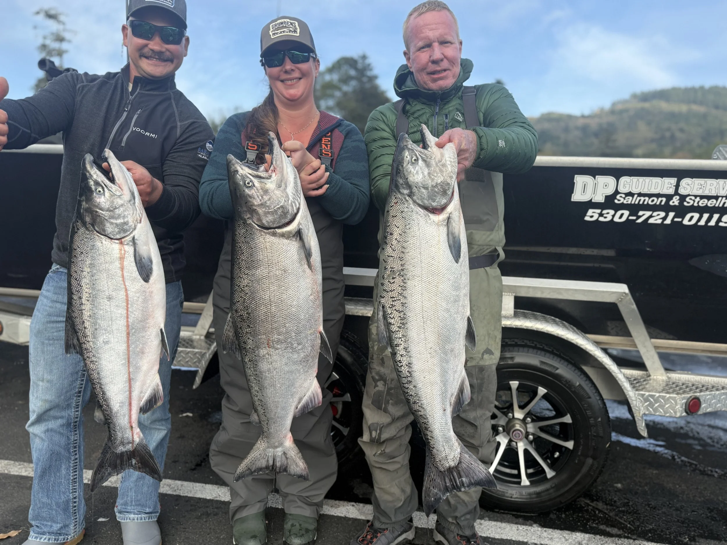 Three people standing in front of a fishing boat, holding large fish they caught. The boat has a sign that reads "DP Guide Service Salmon & Steelhead" with a contact number. The individuals are smiling and dressed in casual outdoor gear.