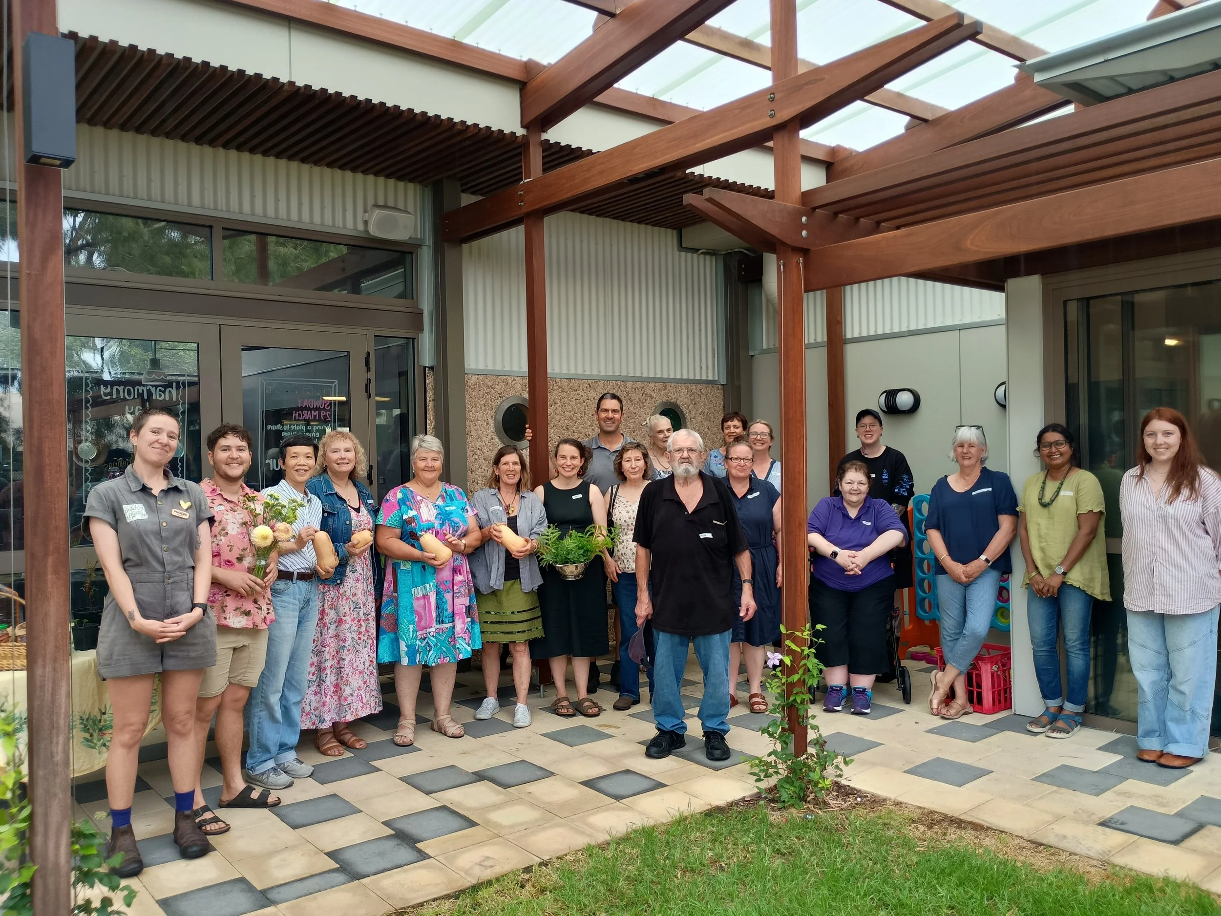 A large and diverse group of people standing outside a building and smiling for the camera. Some are holding vegetables