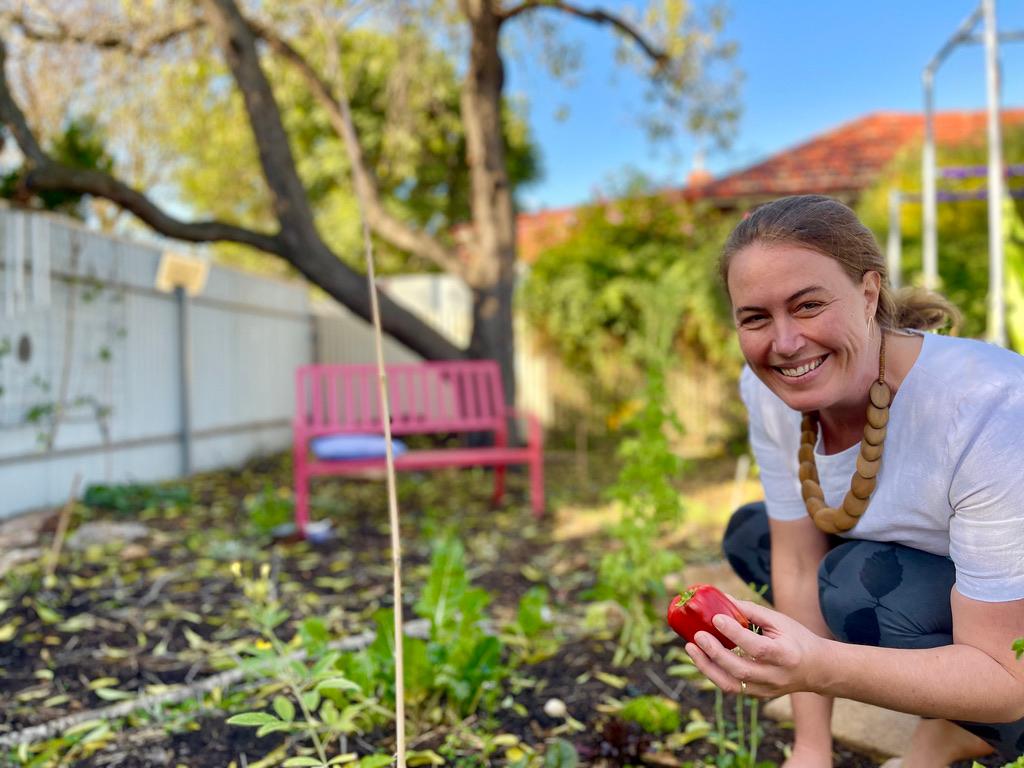 Woman with long light coloured hair kneeling in a garden holding a newly picked red capsicum. She wears dark blue pants, a white t-shirt and a chunky wooden necklace
