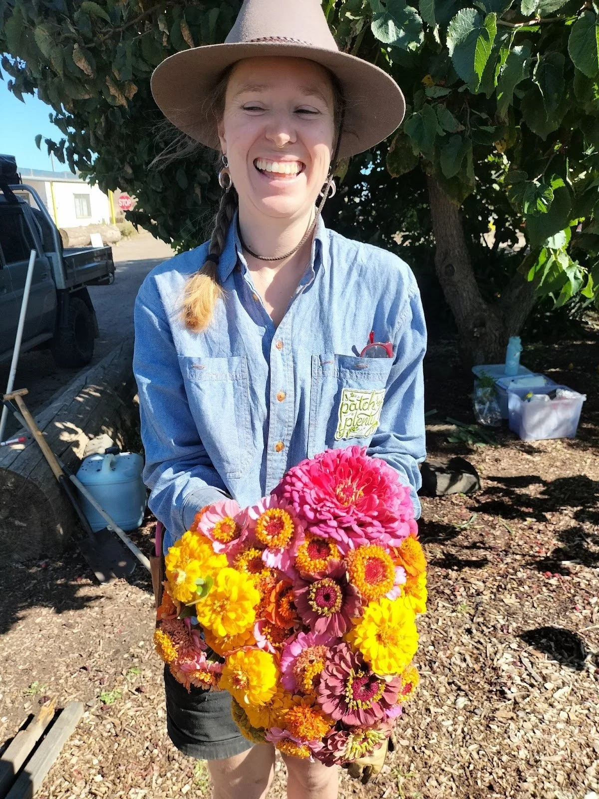 Woman in a light blue collared shirt and an outback hat holding a bright bunch of flowers. She wears her blonde hair in a long braid and is standing in a garden