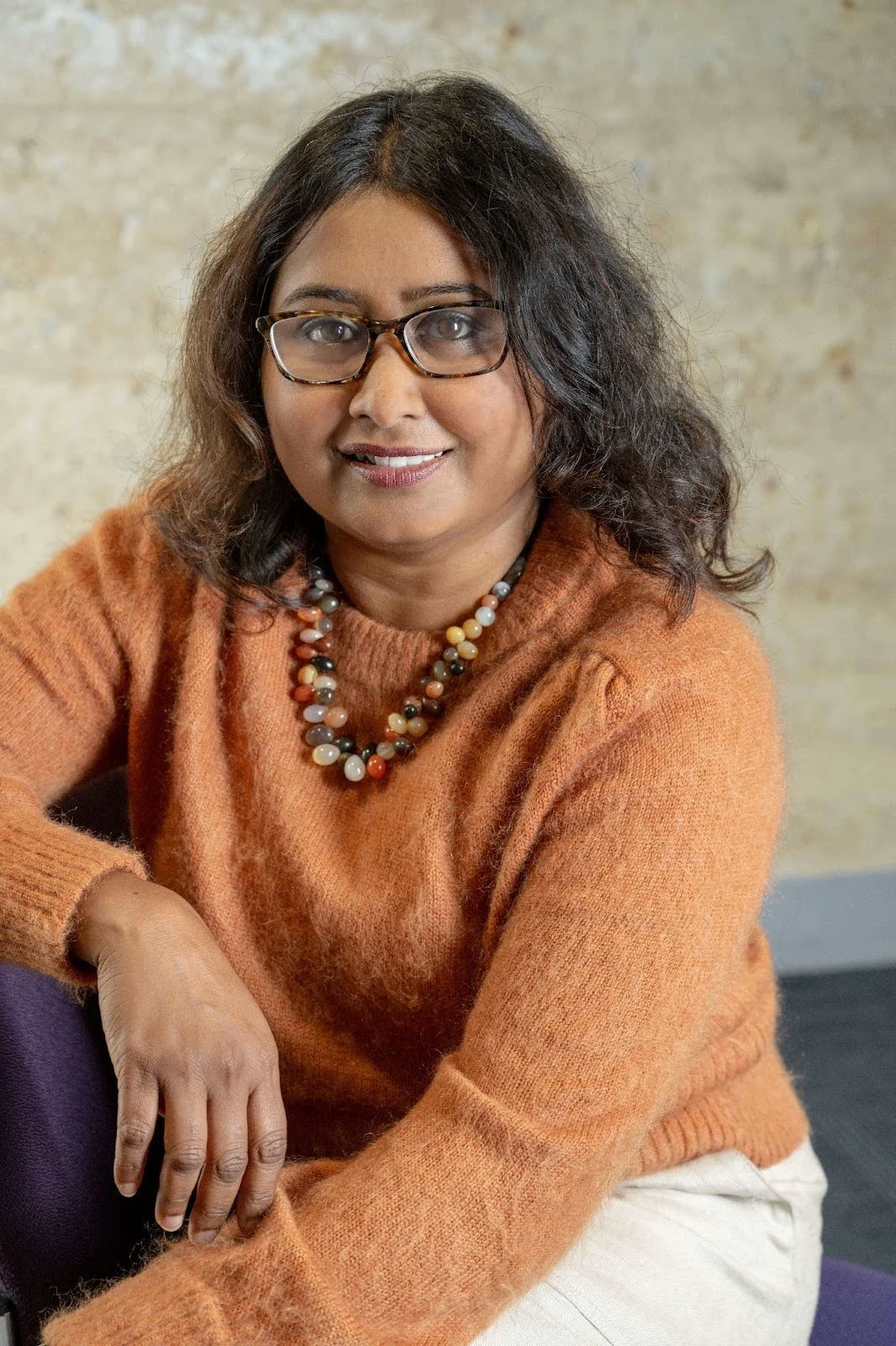 Sultana Razia is wearing an orange jumper and a beaded necklace. She has shoulder-length dark hair and tortoise-shell glasses