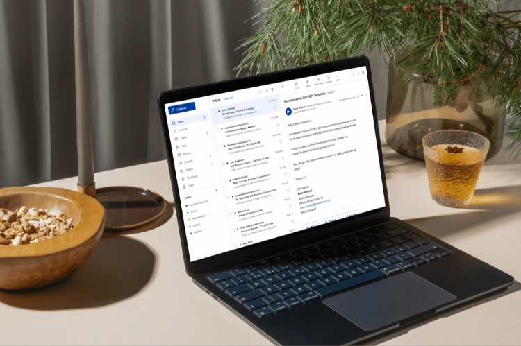 A laptop on a desk displaying an email inbox with open messages, next to a glass of tea with star anise, a wooden bowl with snacks, a lamp, and a potted plant.