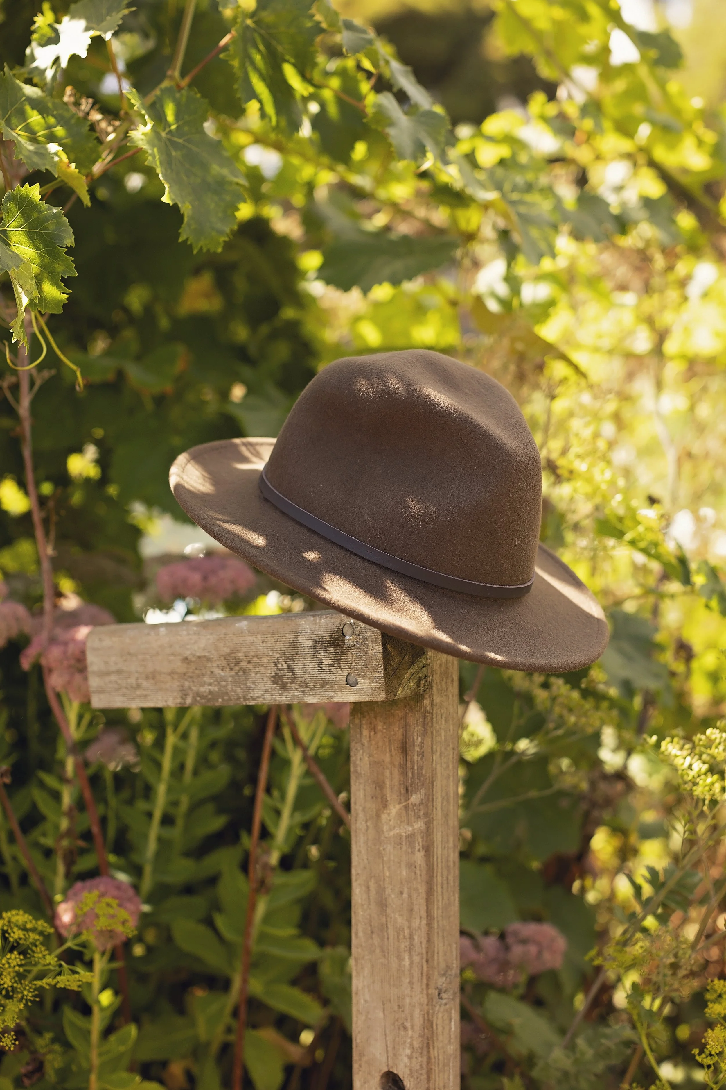A brown wide-brimmed hat resting on a wooden post outdoors, surrounded by green foliage and pink flowers.