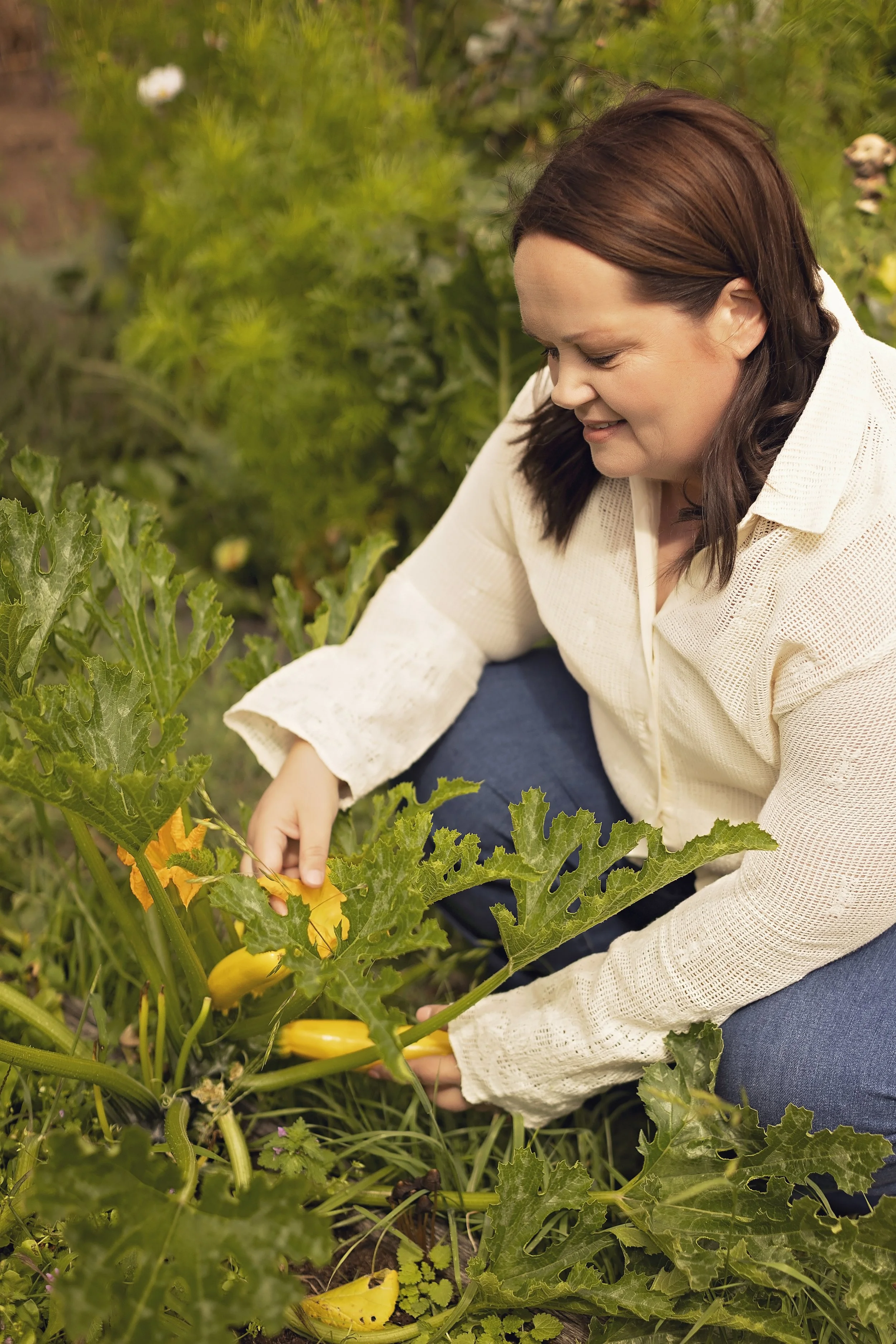 A woman with dark brown hair wearing a cream-colored long sleeve shirt and blue jeans is squatting and picking yellow squash from a garden filled with greenery and flowering plants.