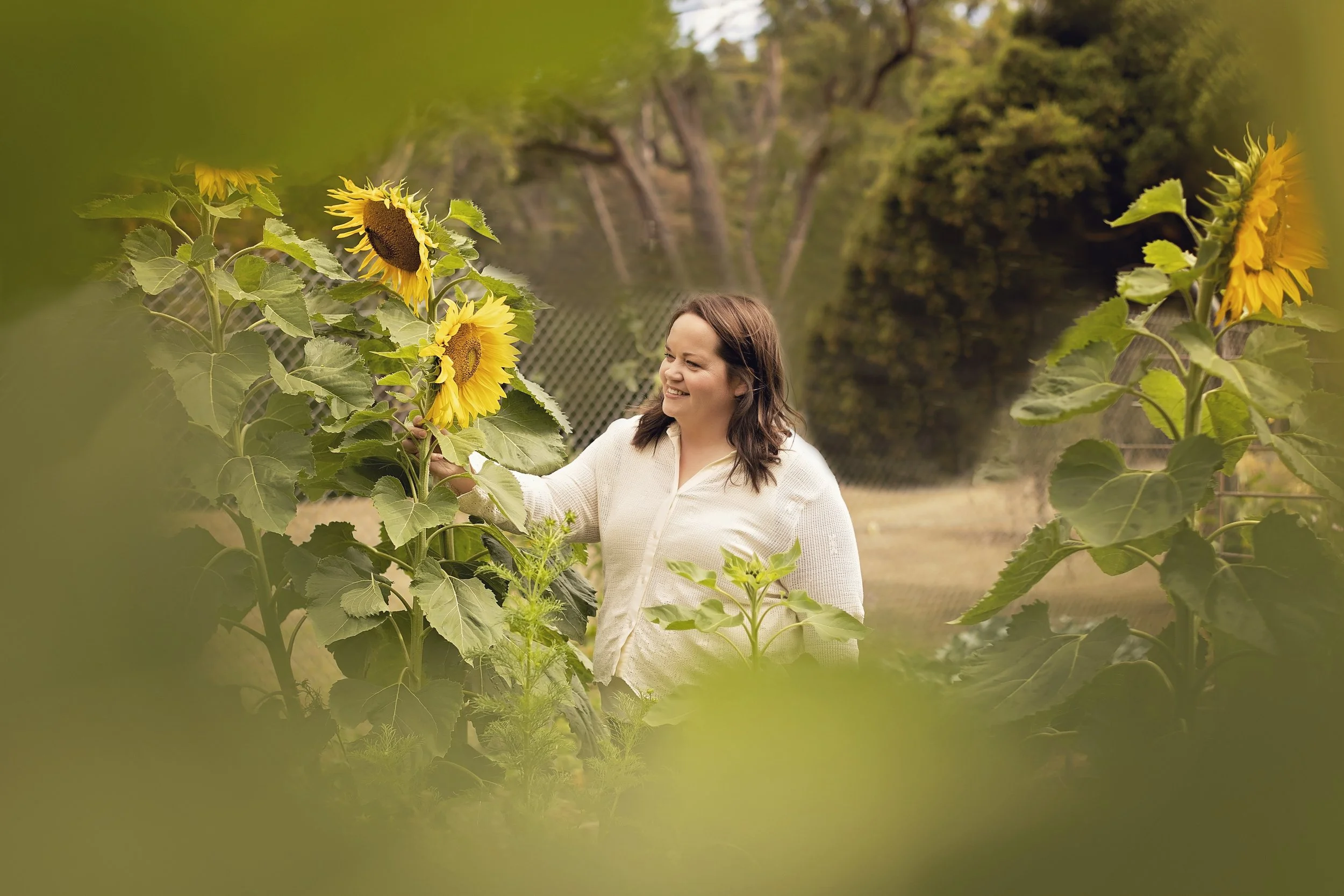 A woman smiling and holding a sunflower in a garden with other sunflowers and greenery around her.