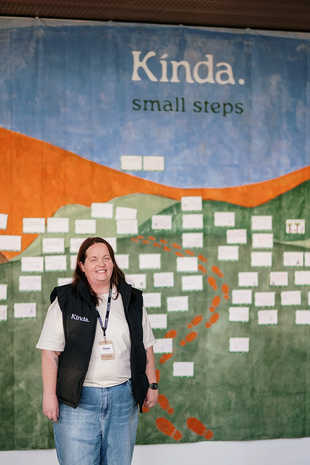 A woman standing in front of a colorful mural with the words 'Kinda. small steps' at the top, wearing a black vest with 'Kinda.' embroidered on it, a white t-shirt, and a name tag that says 'Claire'.