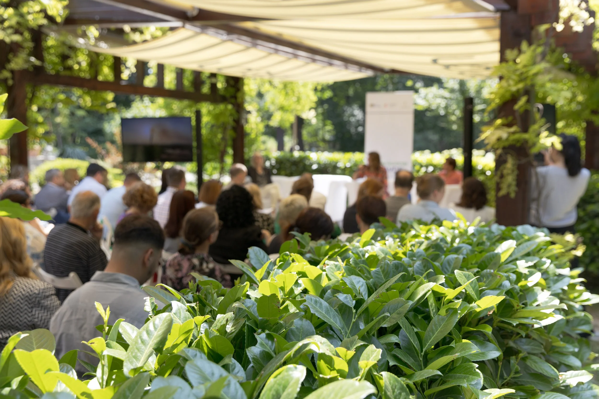 An outdoor gathering with people seated under a canopy, attending a presentation or lecture surrounded by greenery.