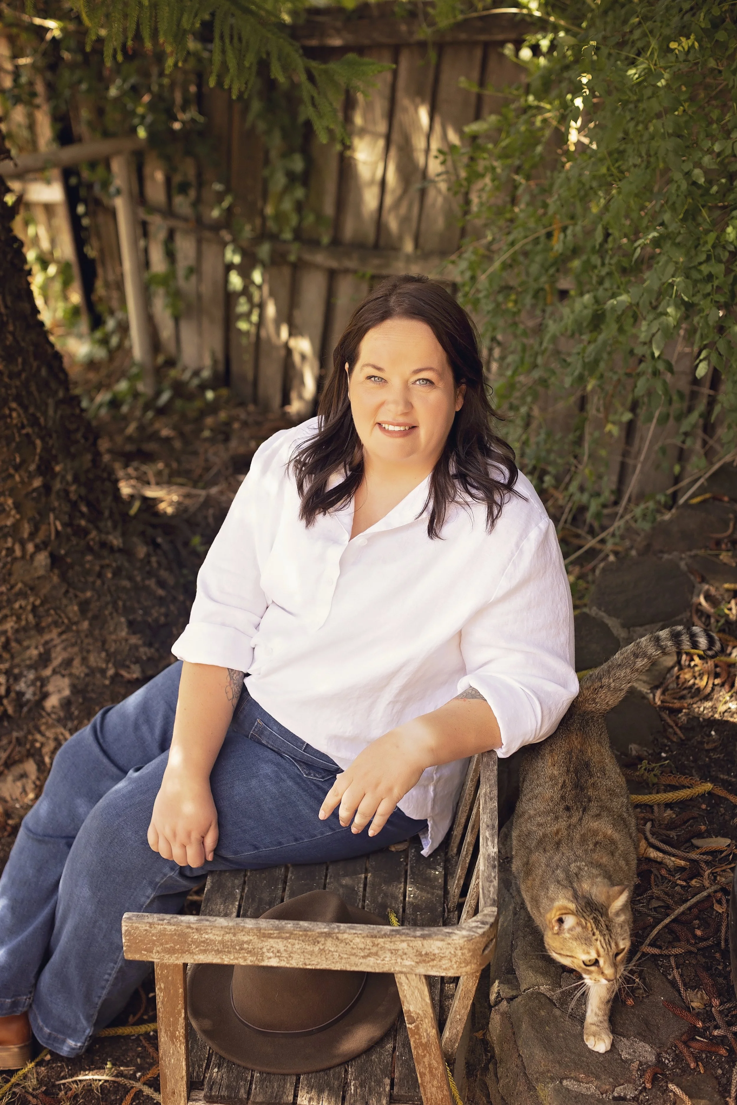 A woman with dark hair sitting on a wooden bench in a garden, with a tabby cat stretching next to her.