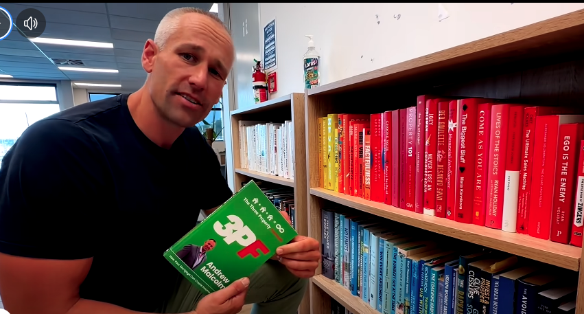 A man holding a copy of the book 'The Prophetic 101' by Andrew Malcolm in a bookstore aisle with rainbow-colored bookshelves.