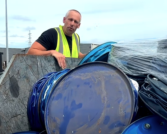 Man in a yellow safety vest looking at blue and black trash barrels and debris outside.