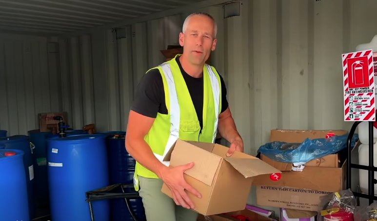 A man wearing a black t-shirt and a yellow safety vest holding a cardboard box in a storage area with blue barrels and a red fire extinguisher sign.