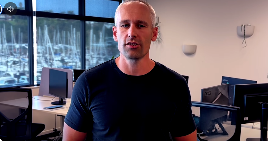 A man in a black T-shirt standing in an office with multiple computer monitors and large windows showing a marina with boats in the background.