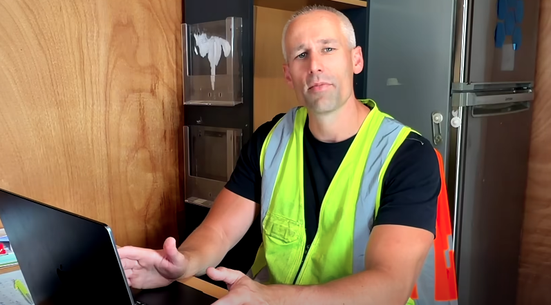 A man wearing a neon safety vest, sitting at a desk with a laptop, in a kitchen with wooden cabinets and a refrigerator in the background.