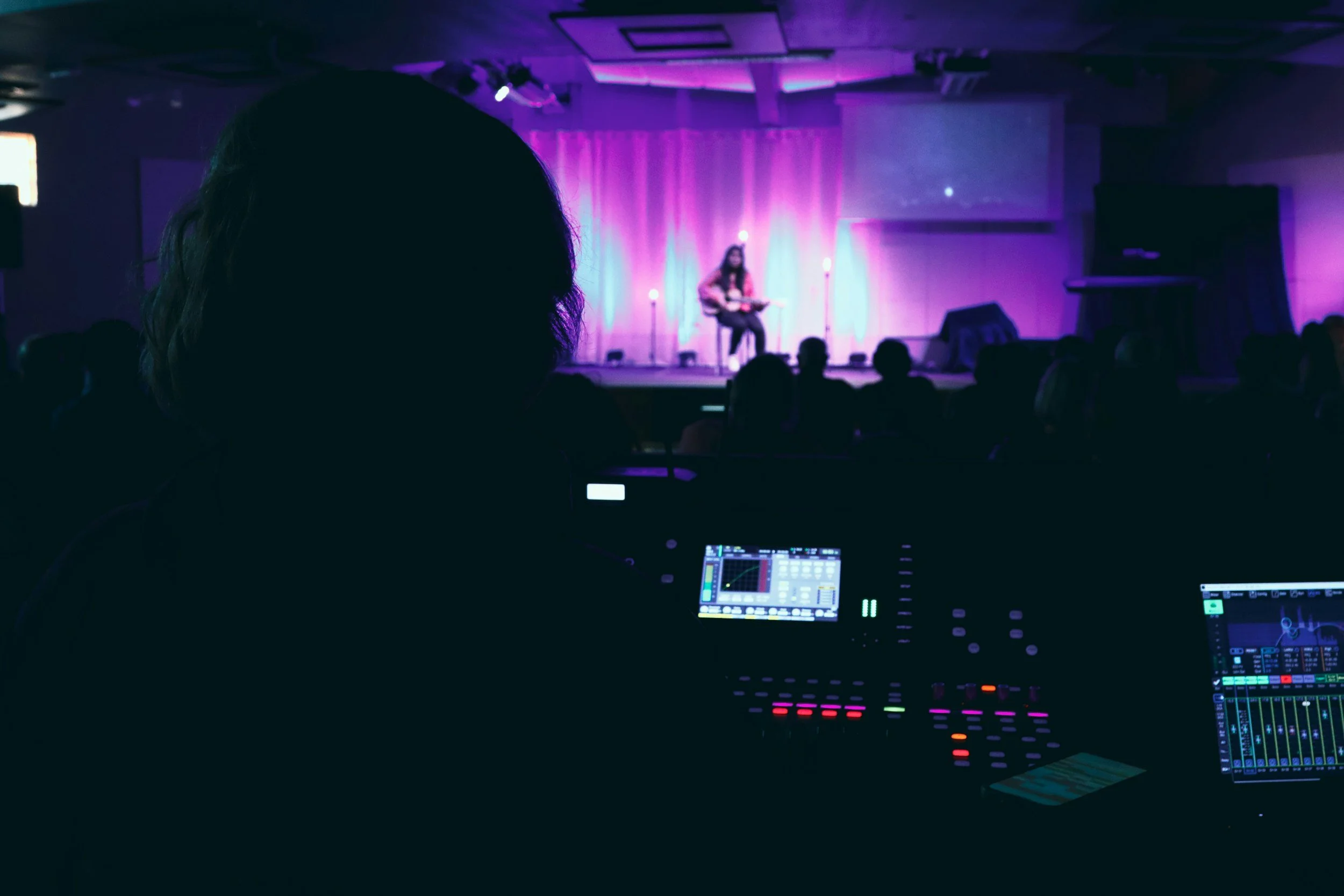 Escenario con cantante tocando guitarra frente a cortina de luces moradas, audiencia en sombras y técnico de sonido controlando la música.
