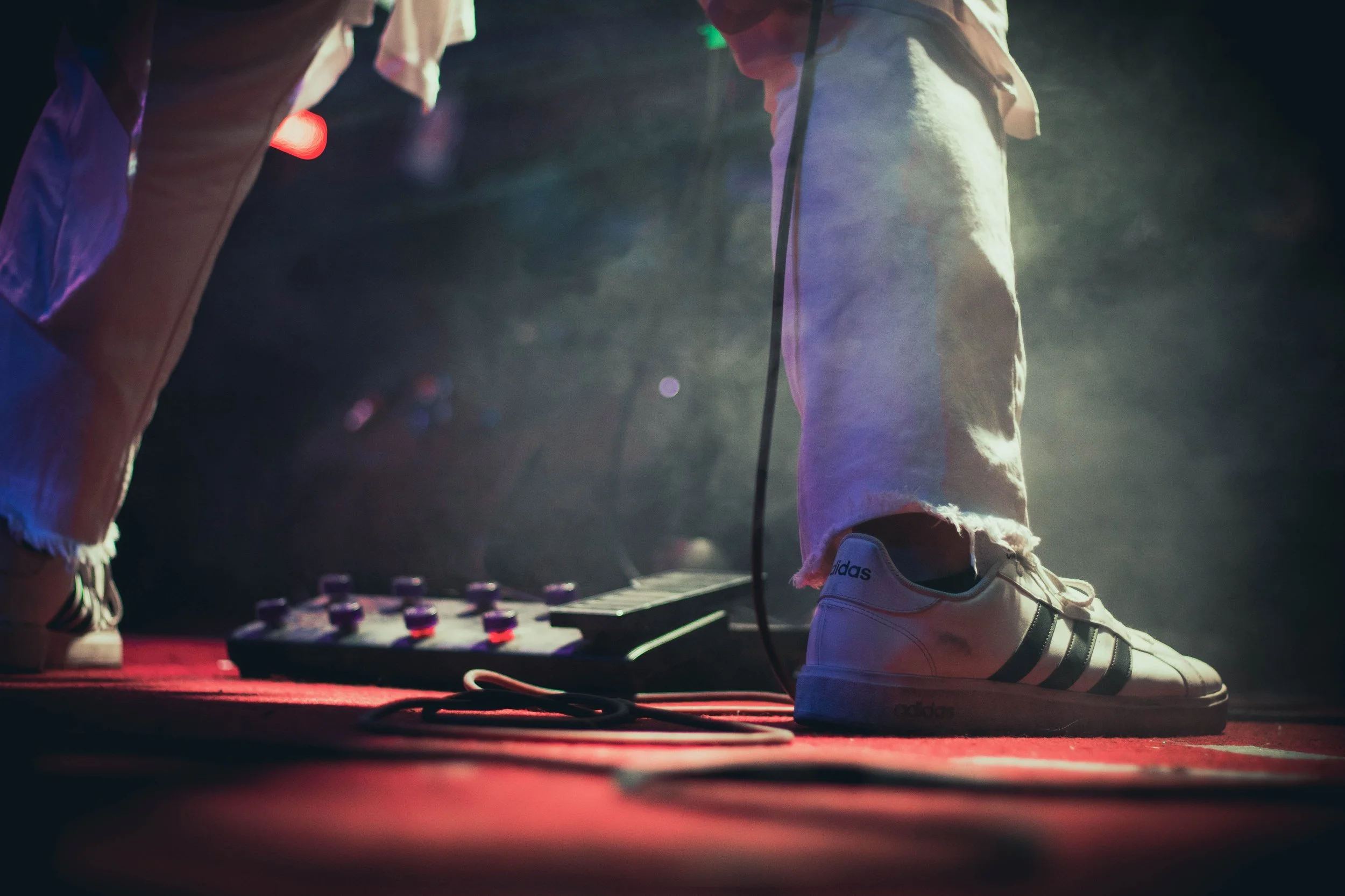 Persona con pantalones rasgados y zapatos deportivos blancos con rayas negras, de pie sobre un escenario con cables y efectos de luz, junto a una mesa con equipo de música.