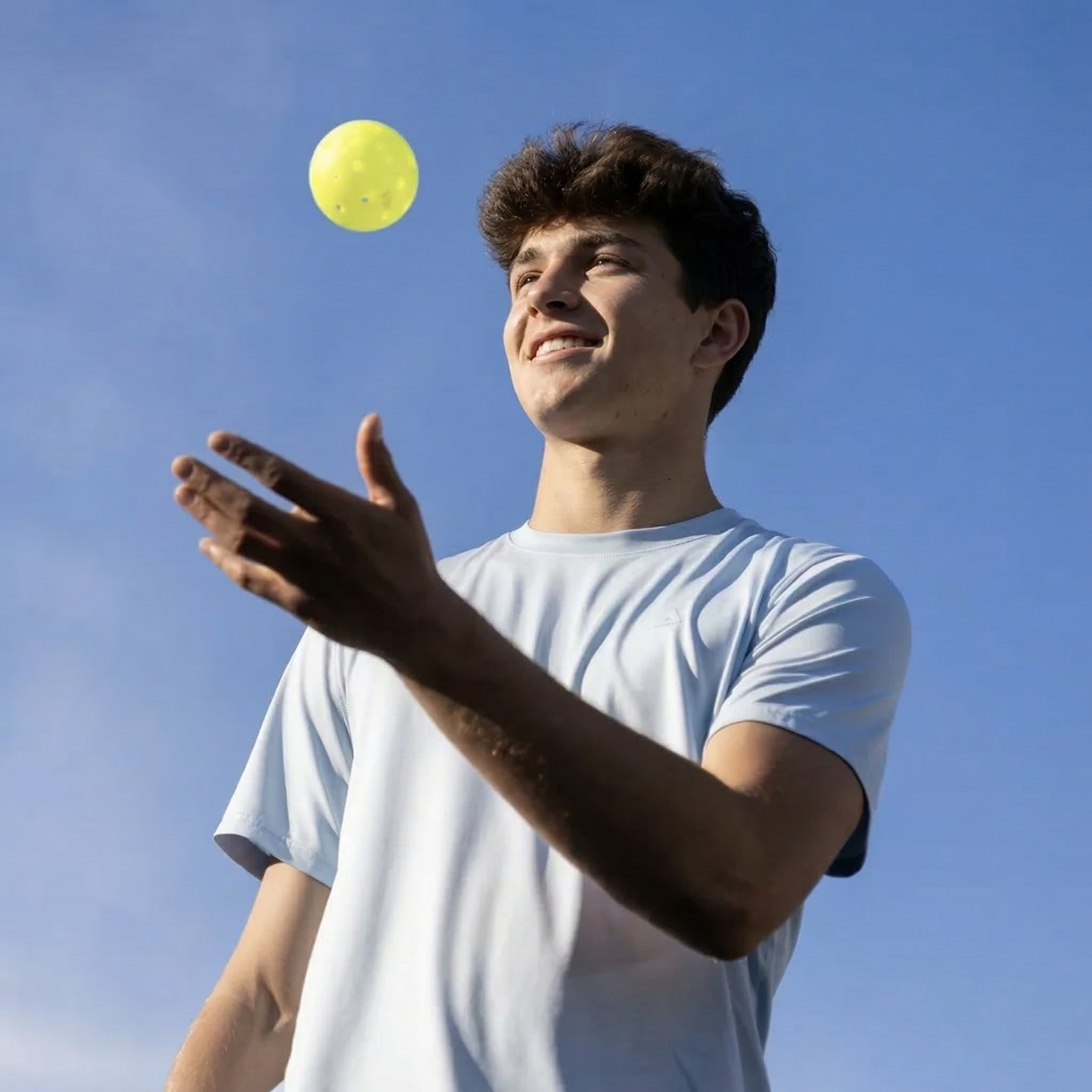 A young man in a white t-shirt tossing a yellow ball in the air outdoors with a clear blue sky in the background.