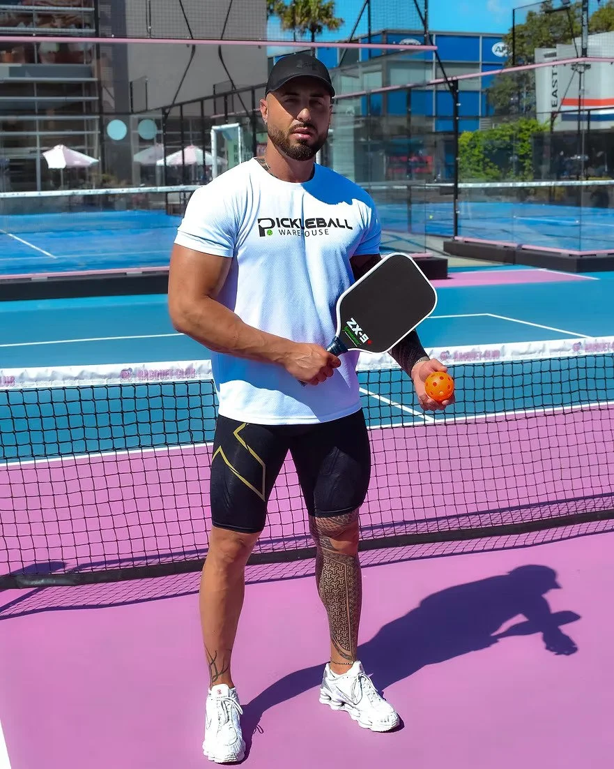 A man standing on a pickleball court holding a paddle and orange pickleball ball, wearing athletic clothing, black cap, and white sneakers, with a net and outdoor sports complex in the background.