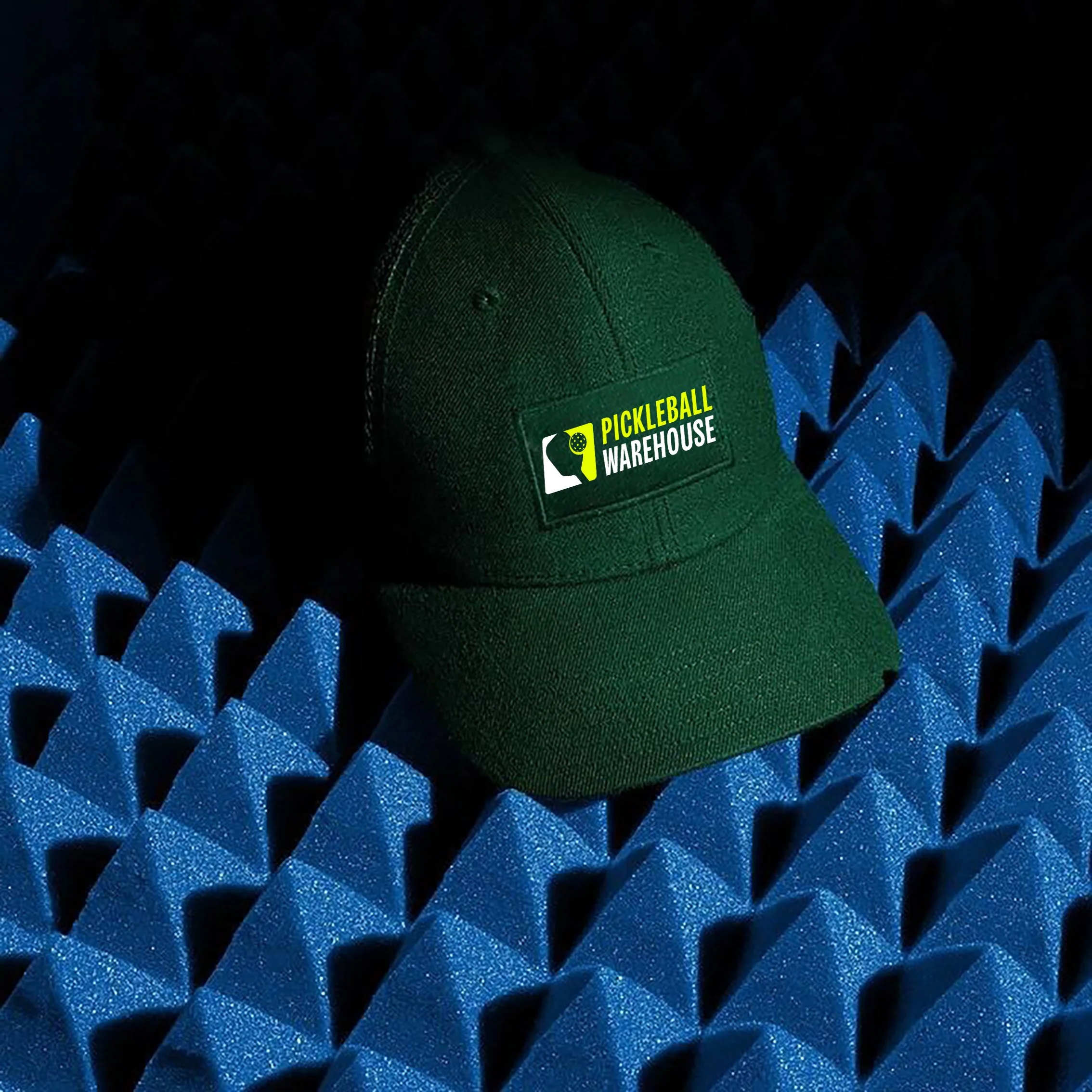 A green baseball cap with a Pickenball Warehouse logo resting on dark blue acoustic foam panels.