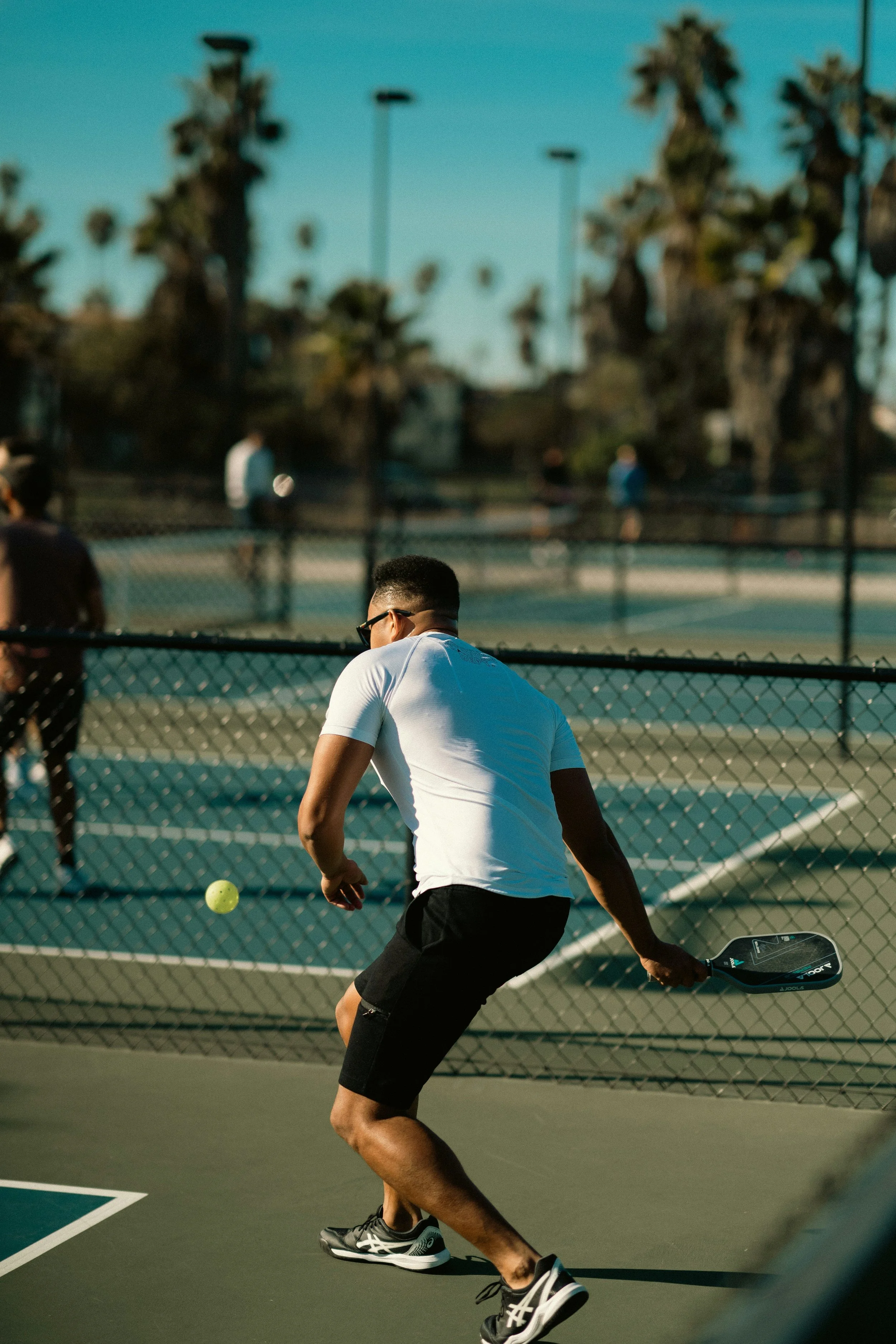 A young man playing pickleball on an outdoor court during daytime, hitting a yellow ball with a paddle, with palm trees and other players visible in the background.