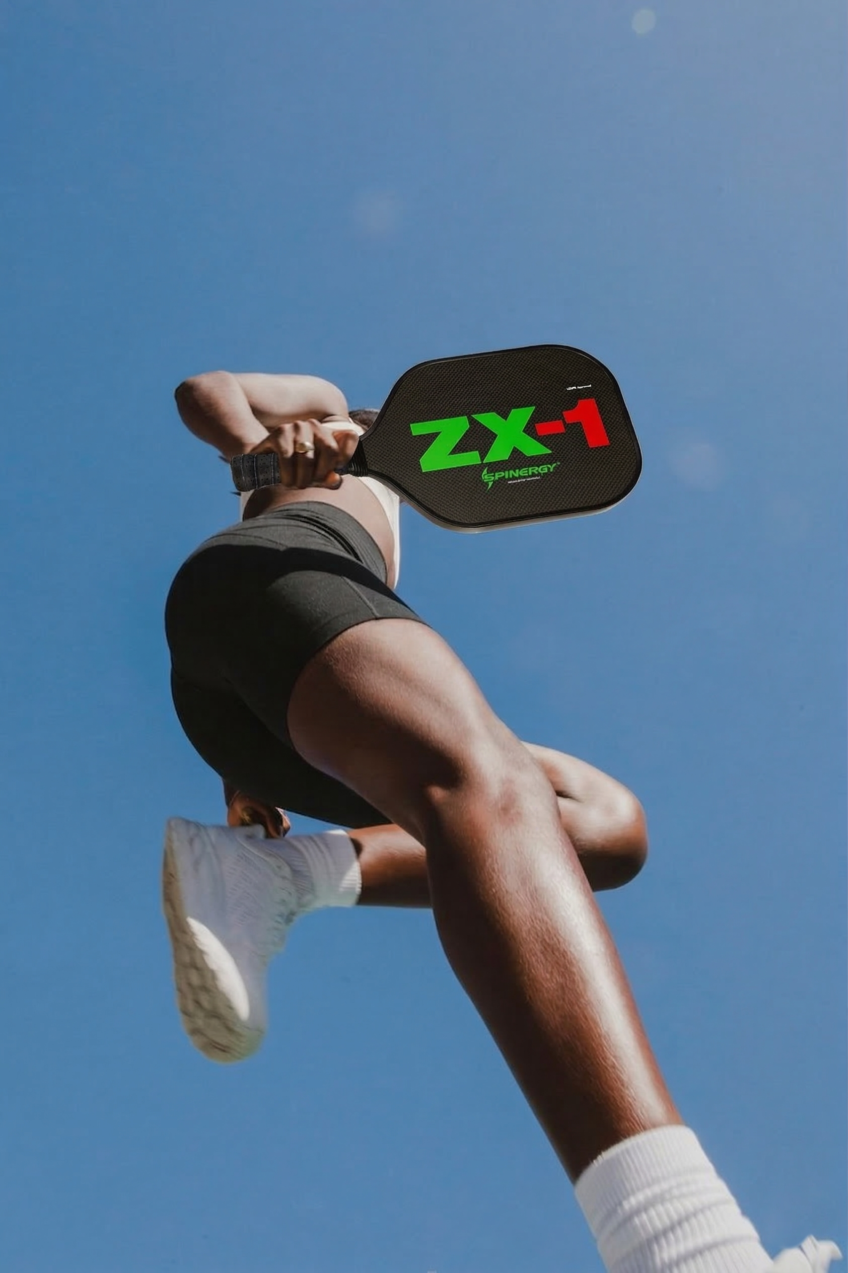 A person in athletic shorts and white sneakers is seen from below, holding a paddle with the text 'ZX-1' and 'SPINERGY' against a clear blue sky.