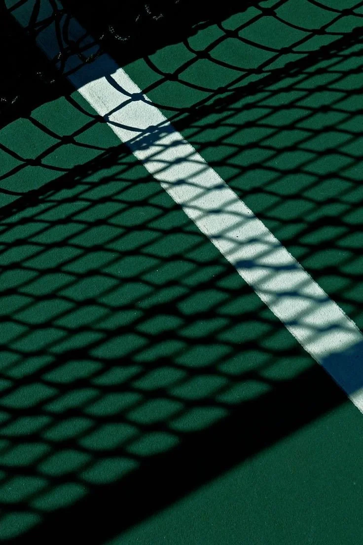 Shadow of a chain-link fence cast onto a green and white tennis court.